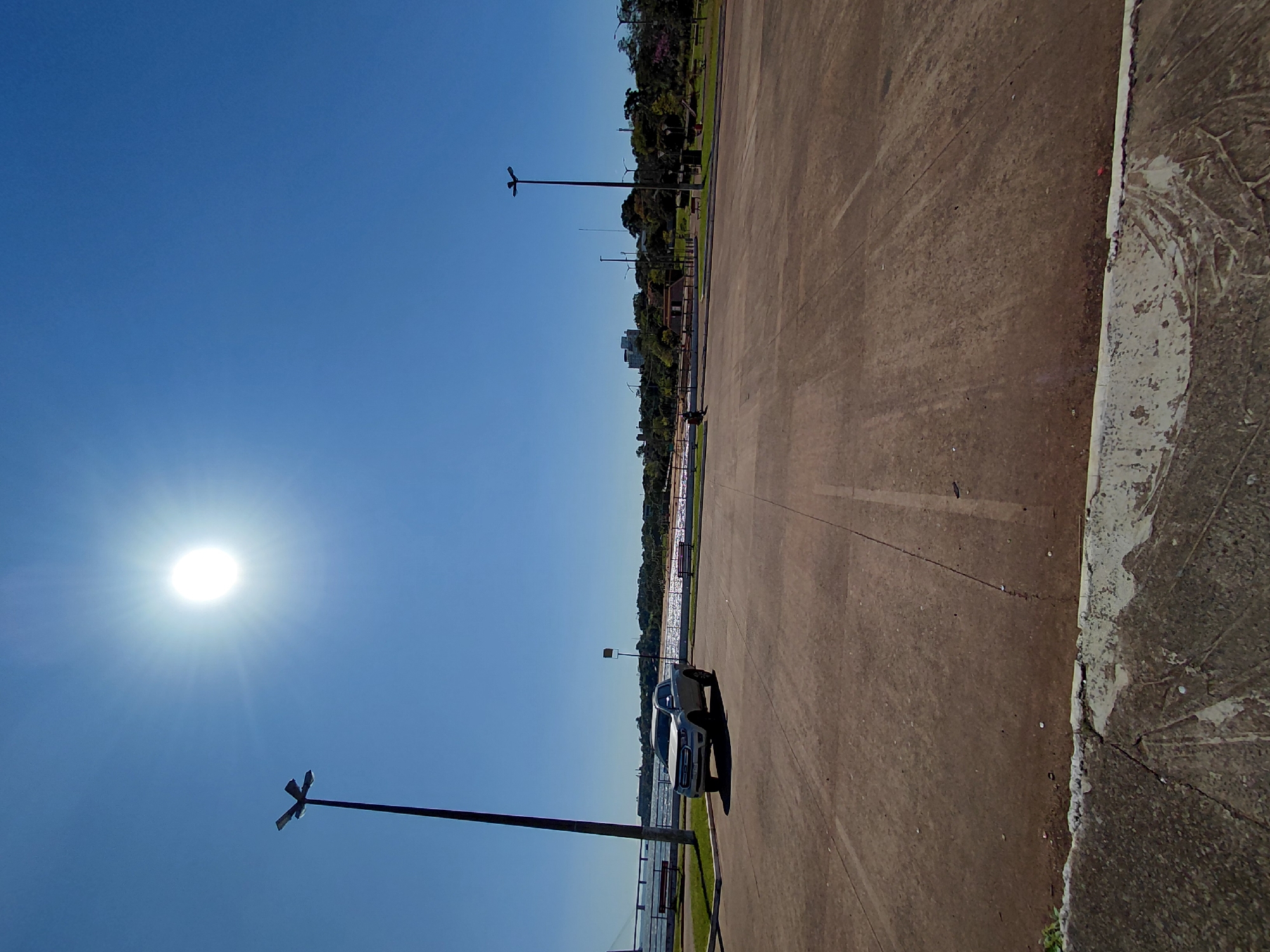 The image depicts an outdoor scene under a clear blue sky with a bright sun. The foreground features a paved surface, likely a parking lot, with visible tire marks and some debris. A light-colored pickup truck is parked on the left side of the paved area. In the middle ground, there is a body of water, possibly a river or bay, with a reflective surface. Beyond the water, a shoreline with a beach and vegetation is visible, leading to a cityscape with buildings in the distance. Several streetlights are present, one tall one prominently on the left and others further back. A bridge structure is partially visible on the far left. The scene is characteristic of the Costanera de Encarnación, a waterfront promenade in Encarnación, Paraguay, known for its recreational areas and views of the Paraná River. There are no people clearly visible in the image.