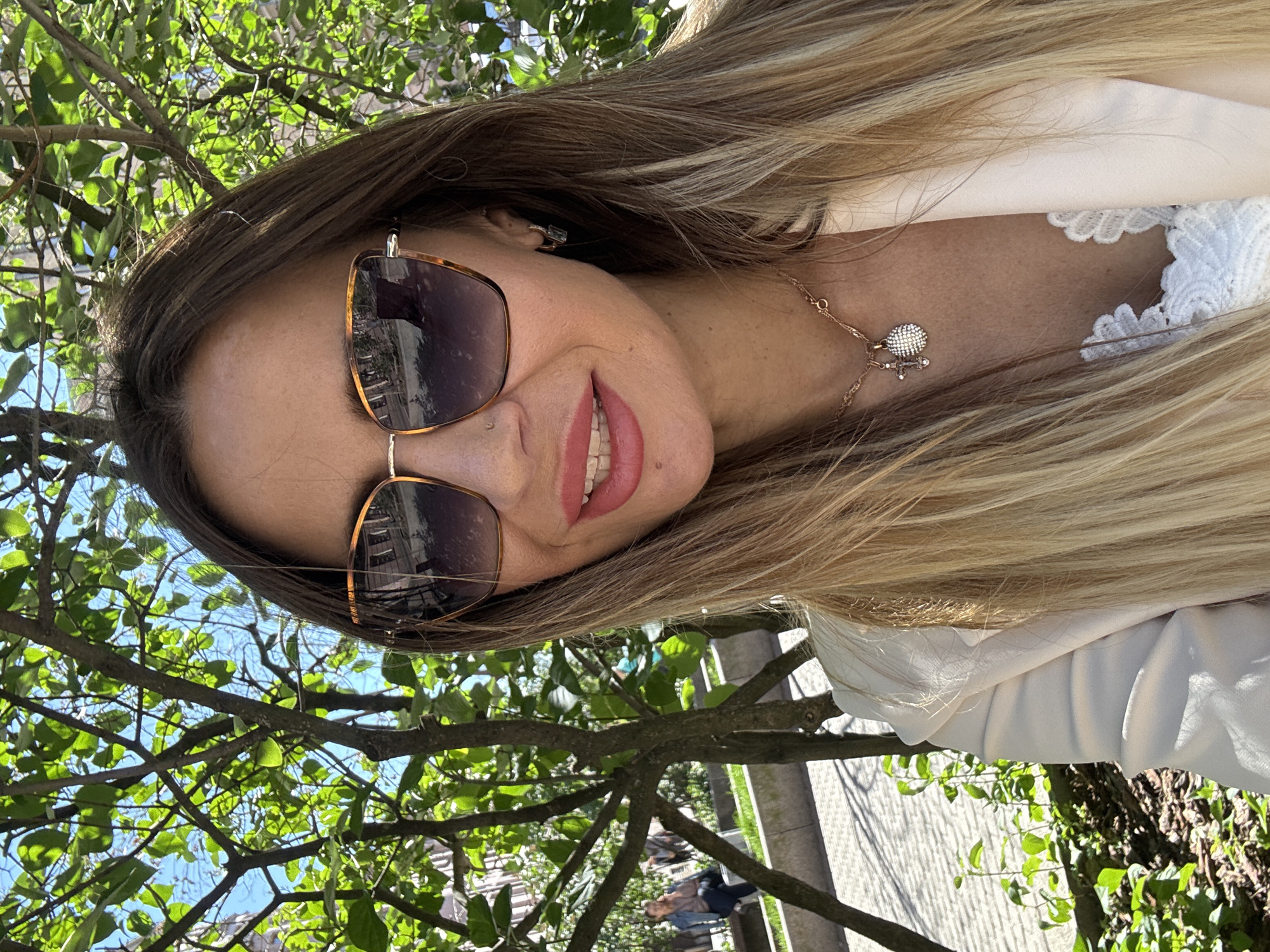 A young woman with blonde hair is taking a selfie outdoors, with trees and sunlight in the background. She is wearing large, tortoiseshell sunglasses and a white jacket over a white lace top. A delicate necklace with a small, sparkling pendant is visible around her neck. Her lips are painted a bright pink, and she has a subtle smile. The image is taken from a low angle, looking up towards the woman, with bright green leaves and blue sky filling much of the frame. The sunlight suggests it is daytime, likely a sunny and pleasant day. In the background, out of focus, are glimpses of buildings and people walking on a paved area, hinting at an urban park or street setting. The overall impression is one of a casual, happy moment captured outdoors.