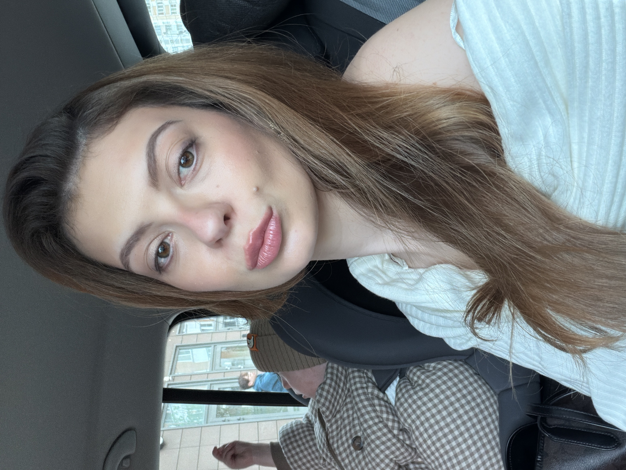 The image shows a woman with long brown hair wearing a white ribbed top in the front passenger seat of a car. Her face is turned toward the camera. In the back seat is a young child wearing a brown plaid jacket and a beanie. The car appears to be stationary with a view of a building with multiple windows visible outside. The location is Khimki, Russia.