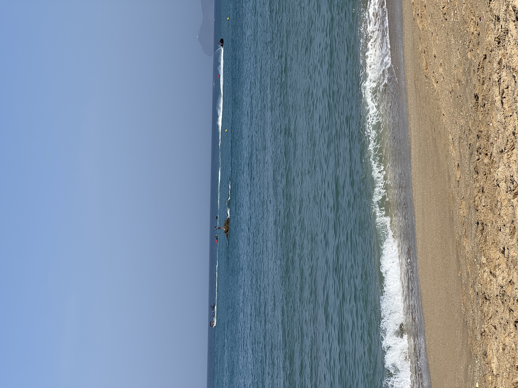 The image depicts a sandy beach with gentle waves lapping the shore. The sea is a deep blue, and in the distance, several jet skis are visible, some creating white wakes as they speed across the water. A small rocky outcrop with a person standing on it is also present in the sea. The sky is clear and blue. The scene is characteristic of a coastal area in M'Diq, Morocco, a popular tourist destination known for its beaches and water sports.