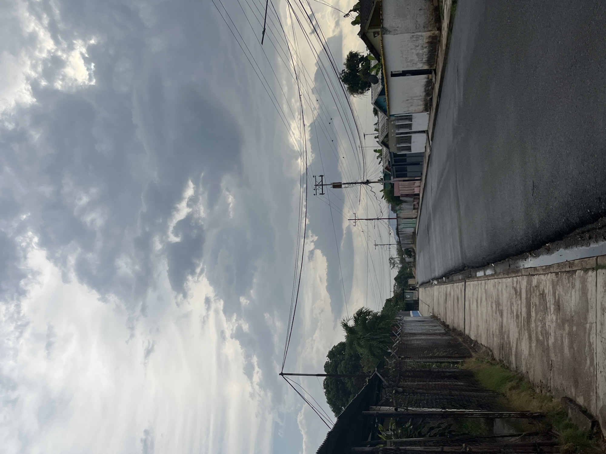 The image shows a street scene in Tinaquillo, Venezuela. The street is wet, suggesting recent rain. The sky is overcast with heavy cloud cover. Overhead power lines are visible. On the right side of the street are buildings, some with visible signs of weathering or age. A sidewalk runs along the edge of the street. On the left, there is a fence and vegetation. The perspective of the photo is from a low angle, slightly tilted. No people are visible in the frame.