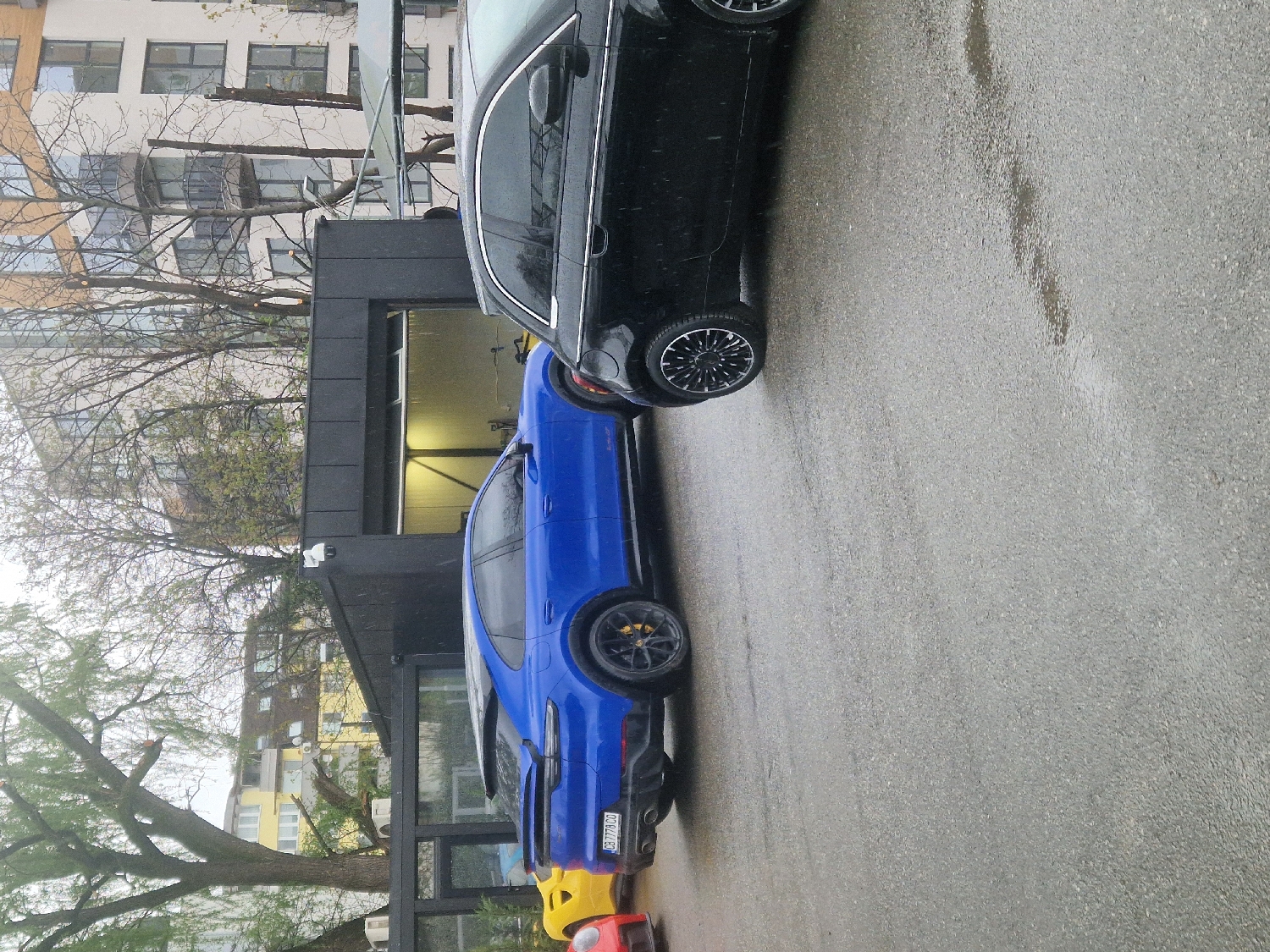 The image, taken in Sofia, Bulgaria, depicts a street scene with several cars parked alongside buildings on a wet asphalt road, suggesting recent rain or damp conditions.
In the foreground, a bright blue car is prominently featured, parked with its rear facing the viewer. It has black multi-spoke wheels with visible yellow brake calipers. Its Bulgarian license plate reads "CB 7782 CO". A portion of a red or orange car with a yellow front bumper is partially visible behind the blue car. In front of the blue car, a black sedan or hatchback is parked, showing its side profile and black multi-spoke wheels.
The road surface is a dark grey asphalt, noticeably wet with reflections and puddles. To the left of the cars, a modern building with dark grey cladding on the lower level and lighter upper stories with numerous windows and some balconies is visible. A security camera is mounted on the dark grey façade. Further in the background, other residential buildings, some with yellow facades and visible air conditioning units, can be seen through the sparse branches of several trees. The trees are mostly bare but show some nascent green leaves, indicating late winter or early spring. The lighting is diffused, suggesting an overcast daytime.