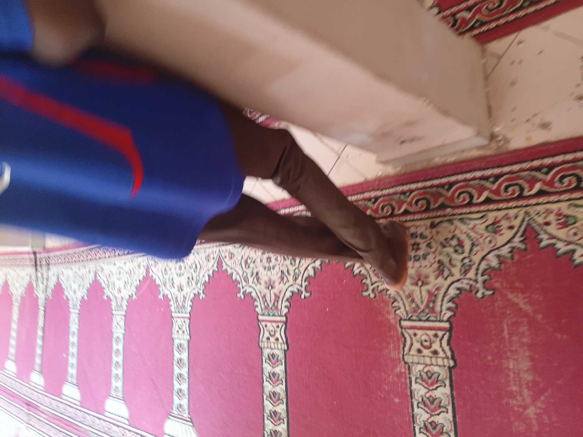 The image shows a low-angle perspective of a person's legs and lower torso, wearing dark trousers and brown shoes, standing on a red carpet with a decorative pattern. The carpet appears to be a prayer mat, indicated by the repeating arched design that is characteristic of Islamic prayer rugs. The person is leaning against a light-colored, possibly concrete pillar or wall. The background is blurred, but suggests an indoor setting with tiled flooring. Based on the presence of prayer mats and the general decor, this scene is likely from a mosque or a prayer room. Considering the location is specified as Jalingo, Nigeria, this would be a mosque in that city. The lighting is moderate, suggesting it's daytime. There are no other people clearly visible in the frame.