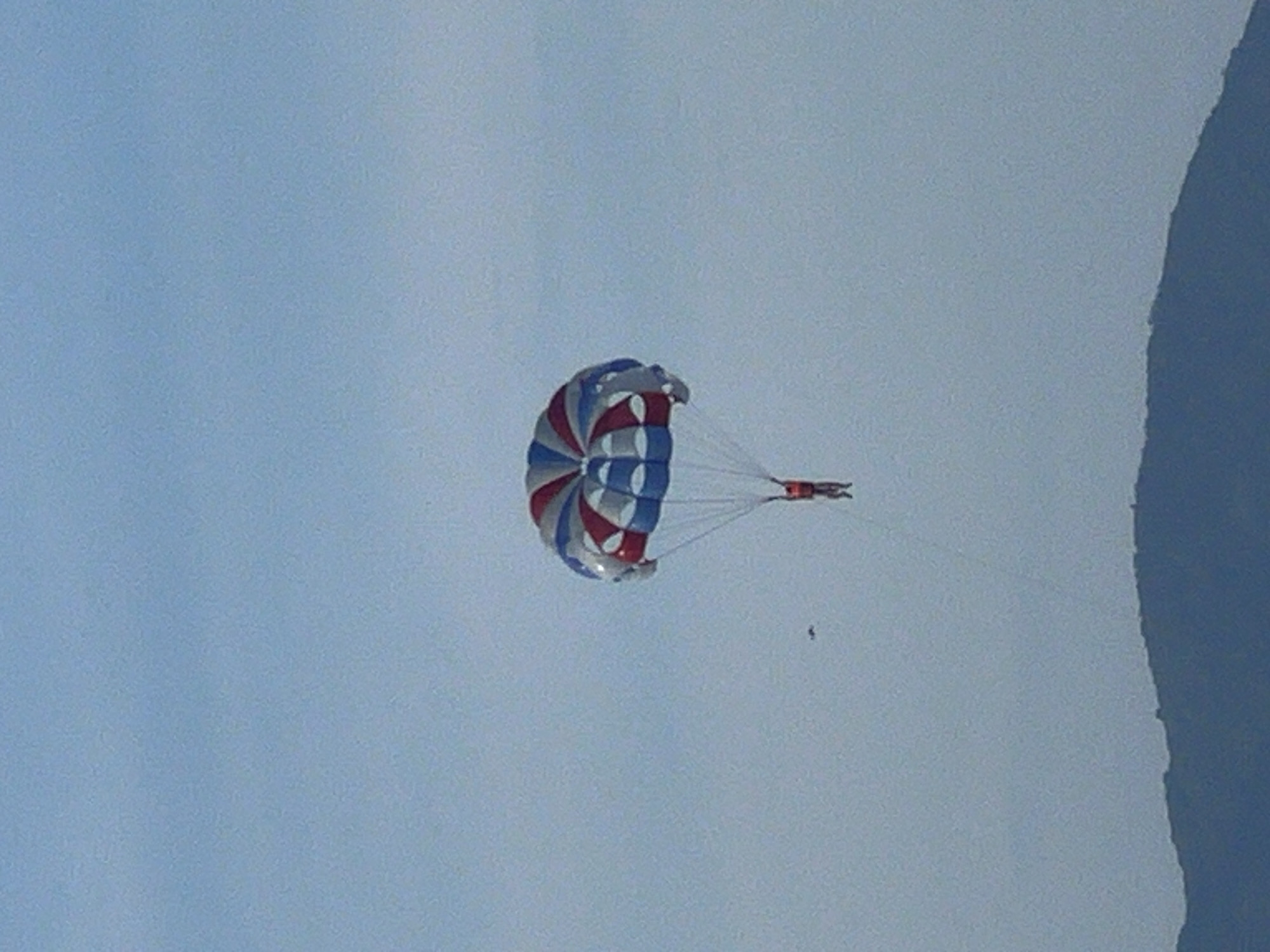 An individual is parasailing against a hazy sky, with a dark, mountainous silhouette in the foreground. The parasail is predominantly red, white, and blue. The person is suspended below the parasail by ropes and is wearing a bright orange life vest. The scene is likely coastal, possibly in Đà Nẵng, Vietnam, given the presence of mountains near a shoreline. The overall lighting suggests it is daytime, but the atmospheric conditions are not clear.