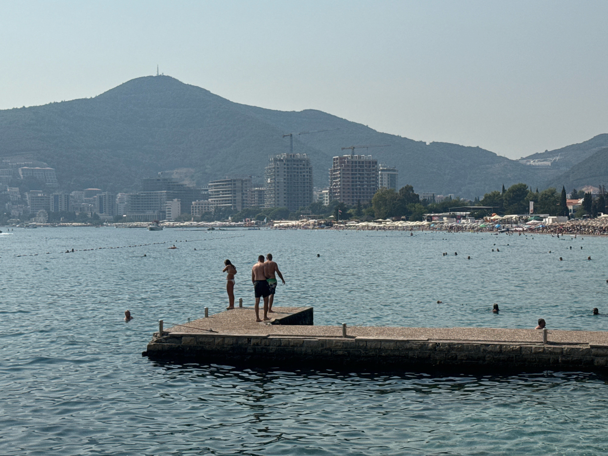 The image depicts a scenic view of the Budva Riviera in Montenegro, likely taken during daytime on a clear day. In the foreground, a concrete pier juts out into the blue, rippling water. Three people are visible on the pier: a woman in a bikini standing to the left, and two men, one wearing swim trunks and the other also in swim trunks, standing close together in the center. The man on the right has his back to the camera.
In the mid-ground, the sea is dotted with numerous swimmers and a few boats. A roped-off swimming area is indicated by buoys. Further back, a bustling beach with many sunbathers, umbrellas, and beach chairs stretches along the coastline. Beyond the beach, a densely populated urban area with modern buildings, some under construction with cranes visible, lines the shore. The backdrop is dominated by a large, tree-covered mountain range, with a prominent transmission tower at its summit. The sky is clear and light blue, suggesting fair weather. The overall impression is one of a lively coastal resort area.