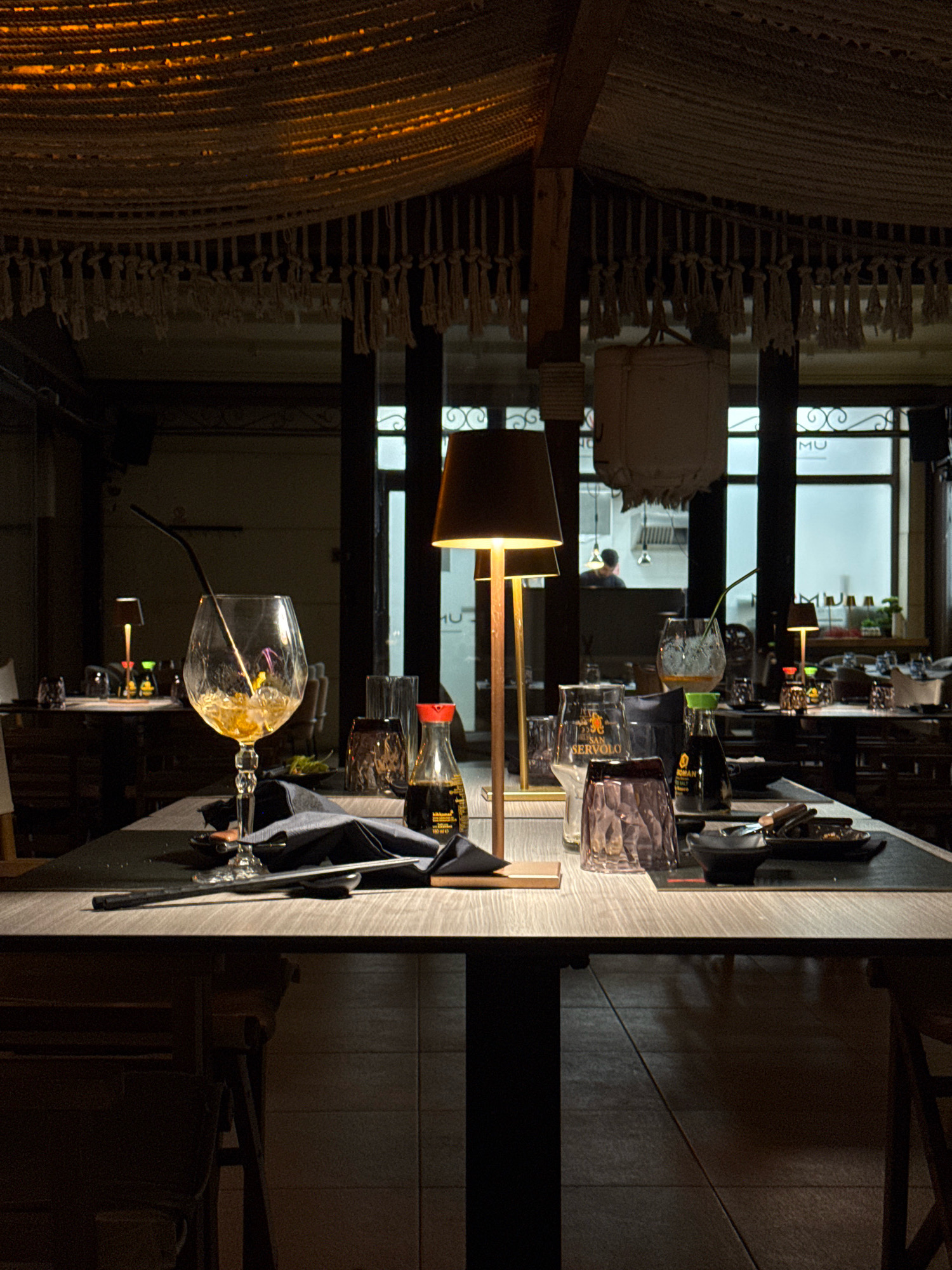 The image depicts a dimly lit restaurant setting, likely in Grad Novigrad, Croatia, with a table set for dining. A large wine glass with a straw and remaining liquid sits prominently in the foreground, alongside a dark napkin and silverware. Other tableware, including tumblers and small bottles, are visible on the table and surrounding surfaces. Two table lamps with warm yellow light provide illumination. In the background, through a window or open doorway, a glimpse of the restaurant's interior can be seen, including other tables and patrons, with the ambient lighting suggesting it is evening. The overall atmosphere is relaxed and intimate, with a focus on the dining experience.