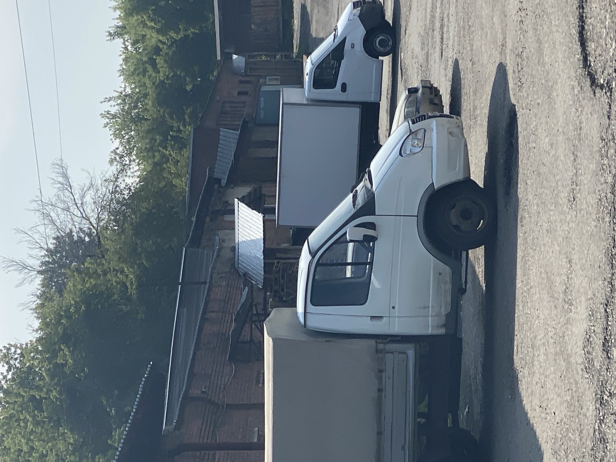 The image shows an outdoor scene with several vehicles parked on a gravel lot. In the foreground, a white truck with a light-colored canvas cover over its cargo area is parked. To its right, another white truck with a closed cargo box is visible. Behind these trucks, a series of brick buildings with metal roofs are situated, and a dense line of green trees forms the background. The sky above is clear and bright. The scene appears to be an industrial or commercial area, possibly a depot or storage facility. There are no people visible in the image. The overall impression is one of a functional, everyday outdoor space.