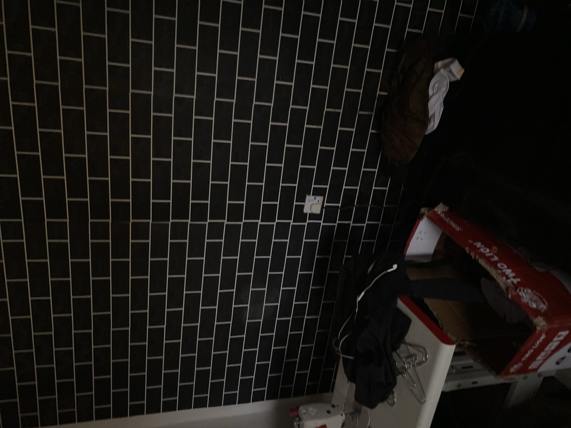 The image depicts an indoor scene, likely a workshop or storage area, characterized by a black tiled wall with white grout. In the lower left quadrant, a white sewing machine is partially visible, suggesting a sewing or tailoring environment. Nearby, a white table holds a pile of dark clothing, some of which appears to be hanging on hangers. To the right of the table, a red cardboard box is visible, displaying Chinese text and graphics, including the words "TWO LION". Inside the box, dark objects are present. Further to the right, on the floor, more dark fabric is strewn about, along with a white cloth with yellow stripes. The lighting is dim, with the tiled wall dominating the background. There are no people visible in the image, and the overall impression is one of a functional, somewhat cluttered space. The location in Jalingo, Nigeria, is not discernible from the visual elements within the image.