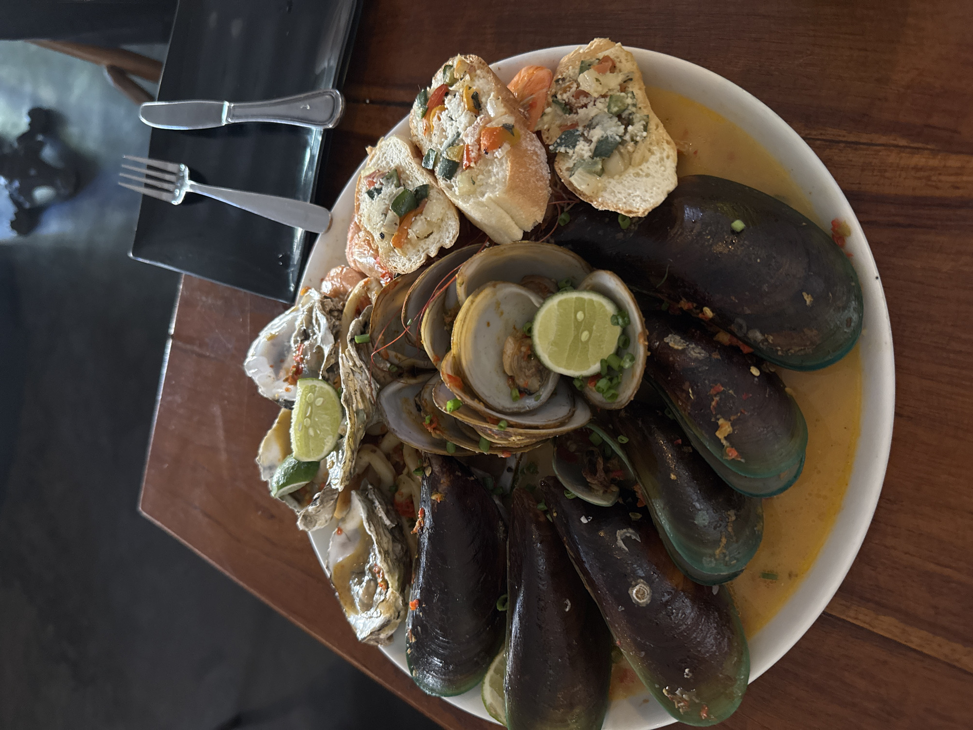An overhead shot captures a platter of seafood served on a wooden table, likely in North Kuta, Indonesia. The platter is filled with mussels, oysters, clams, and shrimp, garnished with lime wedges and chopped scallions. Accompanying the seafood are several slices of toasted baguette topped with a mixture of diced vegetables and cheese. A black plate with a fork and knife sits behind the platter, suggesting a dining setting. The lighting is dim, with a slight glare on the glossy surface of the seafood. The wooden table has a prominent grain pattern, and a dark, out-of-focus background hints at a restaurant interior.