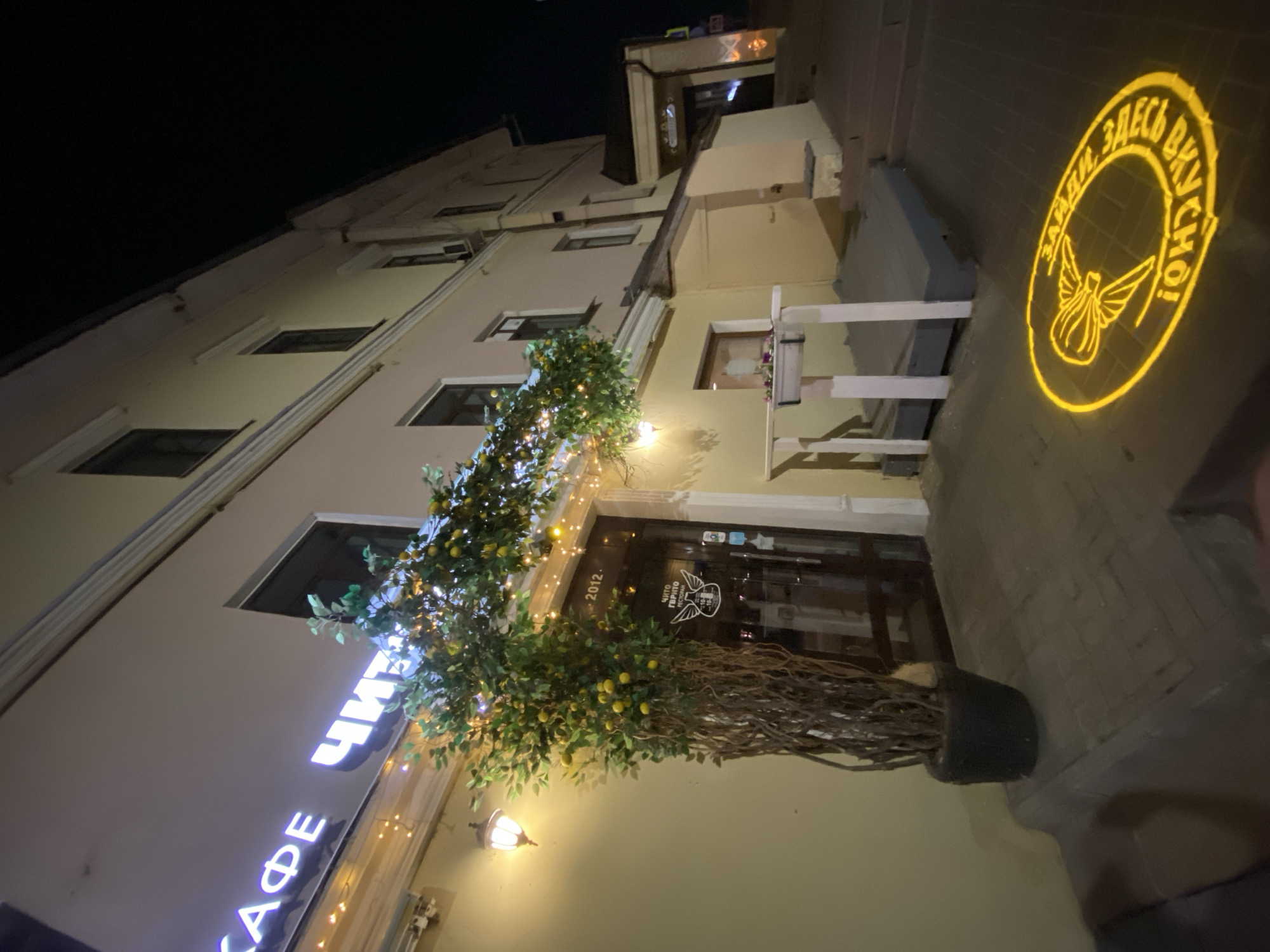 At night, a three-story building in Kazan, Russia is shown. The building is beige with a sign "CAFE ЧИТО" in bright white lettering running vertically along the building's left side. The cafe entrance has a sign on the door that reads "ЧИТО ГЕРМ РЕСТОРАН." A decorative lemon tree stands in front of the entrance. A yellow circular projection on the sidewalk displays an emblem and the Russian text "ЗАЙДИ. ЗДЕСЬ ВКУСНО!"