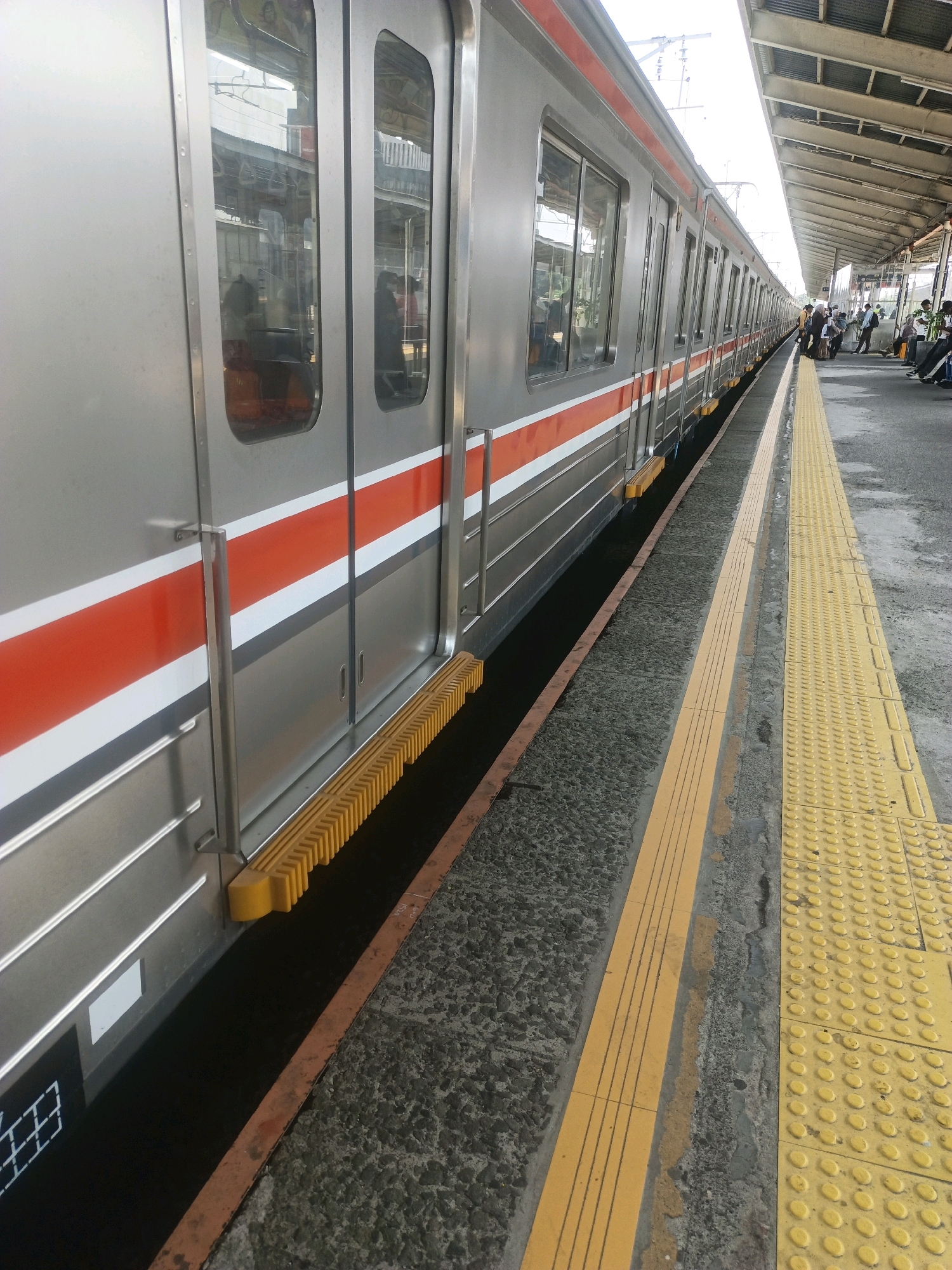The image shows a train at a station platform. The train is silver with red and white stripes along its side. It is a sunny day, and the platform is made of concrete with yellow tactile paving. Several people are visible on the platform, waiting for the train. The station appears to be an outdoor, open-air facility with a roof structure overhead. The overall scene depicts a typical urban train station environment. Based on the image content, the location is likely a train station in Depok, Indonesia.