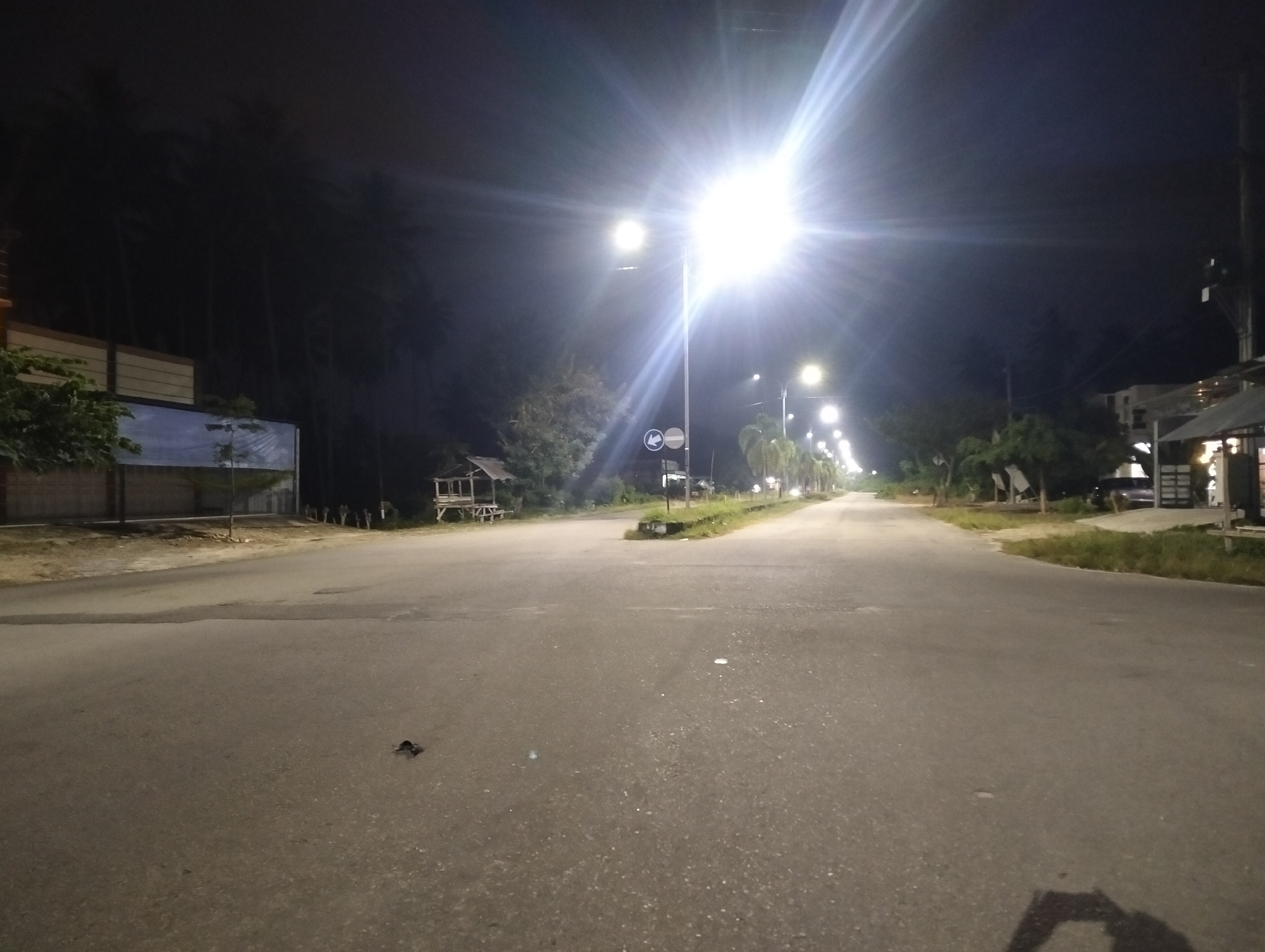 The image depicts a nighttime street scene in Kampal, Indonesia, characterized by artificial lighting from street lamps. The road appears to be asphalt, with some debris visible on its surface. On the left side of the road, there is a structure with a tan facade and a blue mural. Further down the road, a small, open-sided wooden structure resembling a gazebo or shelter is present. Trees line the sides of the road, silhouetted against the dark sky. Multiple street lamps are illuminated, casting bright light and creating lens flares in the image. Some buildings and parked vehicles are visible in the background, suggesting a semi-urban or commercial area. The overall atmosphere is dark, with the lighting drawing attention to the road and its immediate surroundings.