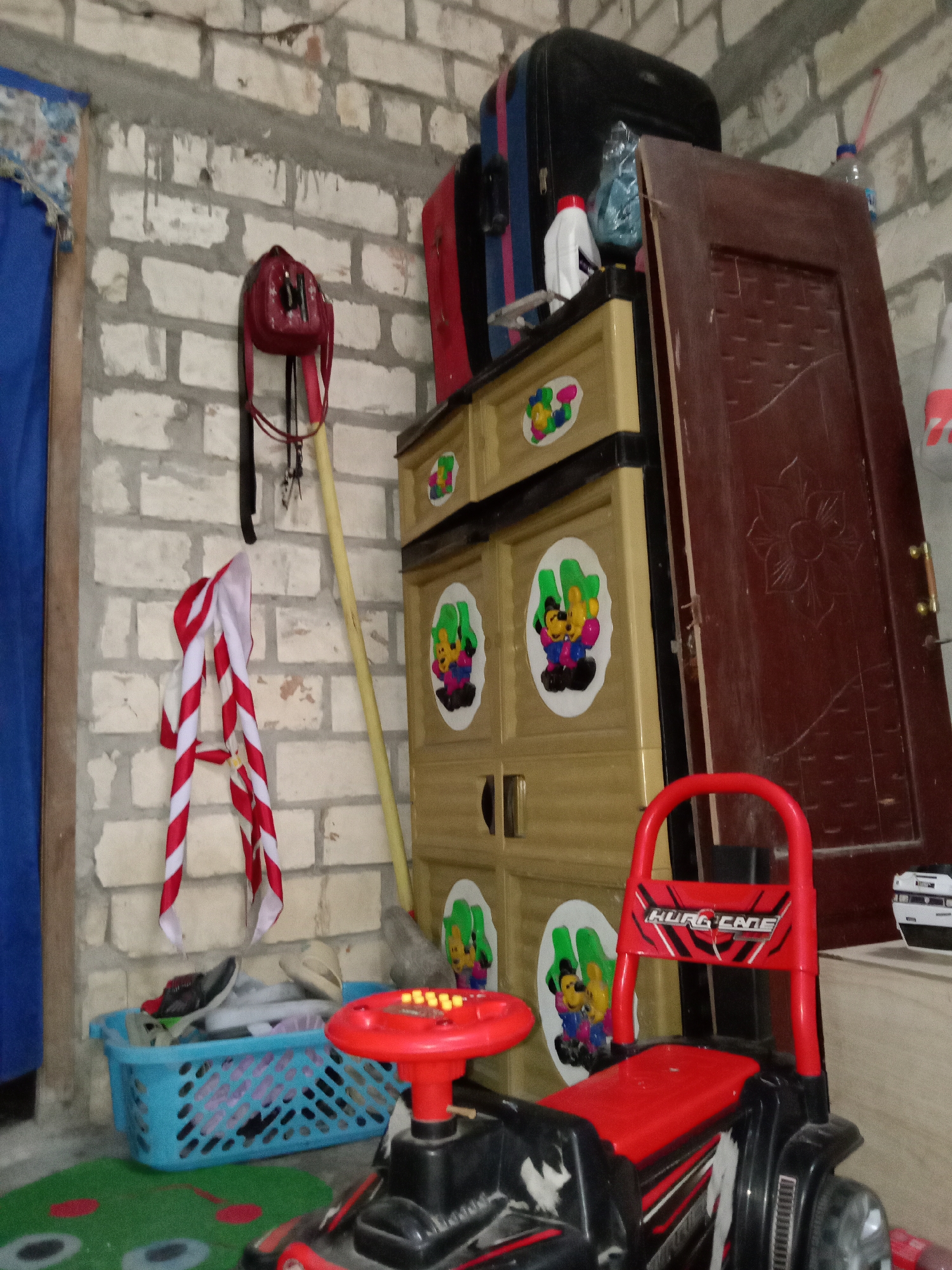The image depicts an indoor scene, likely a storage area or a children's play space, with a rough brick wall as the backdrop. Dominating the center is a yellow plastic cabinet with multiple drawers, adorned with cartoon stickers of characters resembling Mickey Mouse. To the right of the cabinet, a dark red wooden door leans against the wall. On top of the cabinet, various items are stacked, including a black suitcase, a smaller red suitcase, a white bottle with a red cap, and a crumpled blue plastic bag.
To the left of the cabinet, a red handbag with a flower design hangs on the wall, attached to a dark strap. Below this, a red and white striped scarf or bandana is draped. A long, pale yellow pole stands vertically between the scarf and the cabinet, leaning against the wall. In the foreground, a blue plastic basket holds several pairs of shoes, including sandals and sneakers. A small red toy car with a steering wheel featuring illuminated buttons is positioned in front of the basket. The car has the word "HURC GADE" printed on its backrest. A green mat with red circular patterns is visible on the floor in the lower left corner. The overall impression is one of clutter and a lived-in space. The location in the city of Jumput, Indonesia, is not visually evident from the image itself.