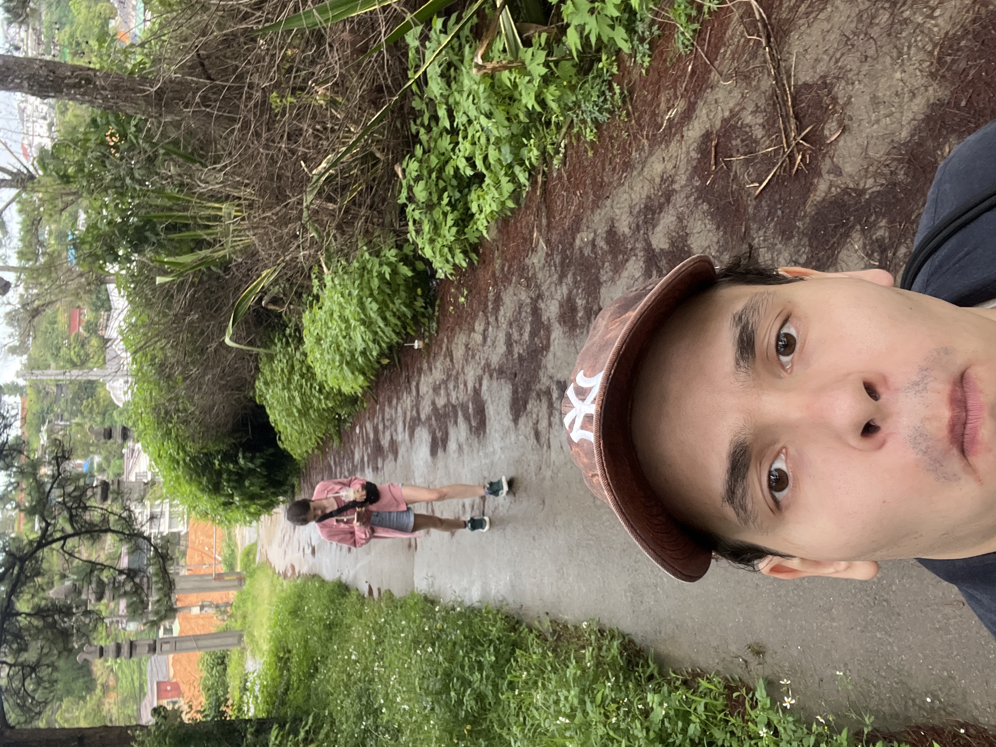 In the city of Cam Ly - Đà Lạt, Vietnam, a selfie is taken from a low angle, focusing on the foreground face of a person wearing a brown baseball cap with a white NY logo. The person's gaze is directed towards the camera. Behind them, a narrow, wet paved path meanders through lush green vegetation. Further down the path, a young woman walks away from the camera, holding a drink in her right hand and looking at her phone. She is wearing a pink jacket, a grey skirt, and dark sneakers. The surroundings feature dense foliage, trees, and what appear to be stone pillars and buildings in the background. The overall atmosphere suggests an overcast or recently rained-upon environment.