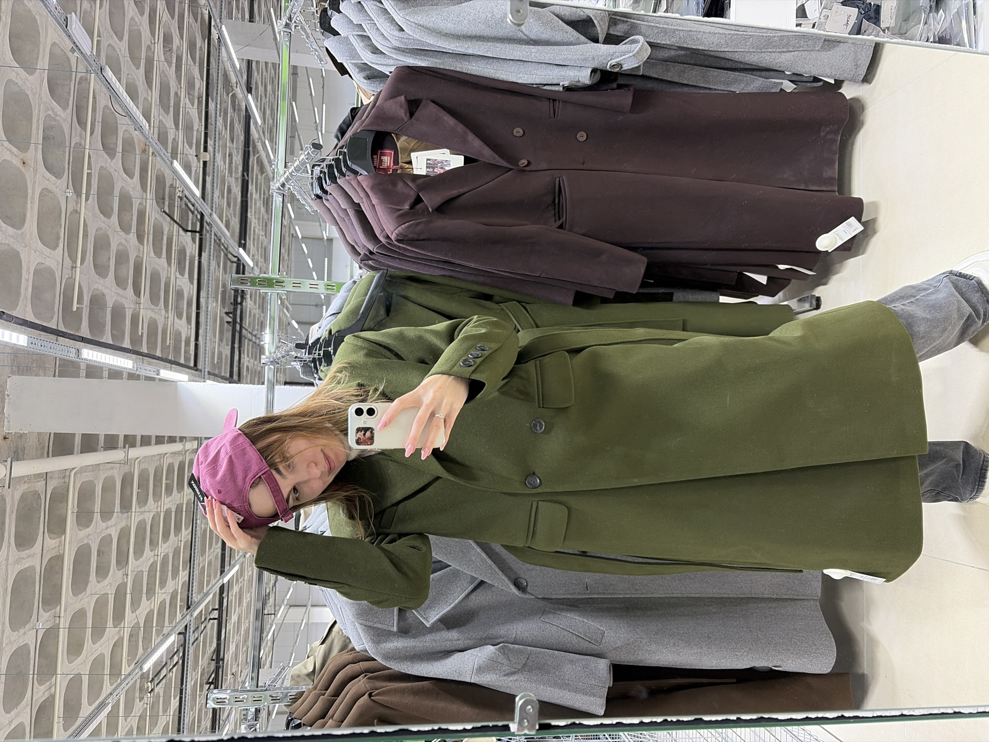A young woman is captured in a mirror selfie, standing upright in what appears to be a retail store or fitting room. She has long, light brown hair and is wearing a dark olive green long coat with a belt, blue jeans, and a pink baseball cap. She holds a white smartphone with two camera lenses in her right hand to take the photo, while her left hand rests on the brim of her cap. A ring is visible on her left ring finger. Her expression is calm and neutral, with a slight smile.
The setting reveals rows of clothing racks behind her, laden with coats in various shades, including grey, brown, and olive green, suggesting a fashion retail environment. Some coats have tags; one brown coat has a tag that appears to say "Leopoldi" or "Leopold", and another label visible on a stack of coats reads "next". The woman's pink cap has a small black tag on the side that faintly reads "KOREA".
The background, visible in the mirror's reflection, features a concrete wall with a distinctive grid pattern of recessed, oval-shaped indentations. Long, linear fluorescent light fixtures are mounted on both the ceiling and along the walls, providing artificial illumination. The overall scene indicates an indoor setting, likely during daytime, given the commercial nature of the space. The image is taken in Izhevsk, Russia.