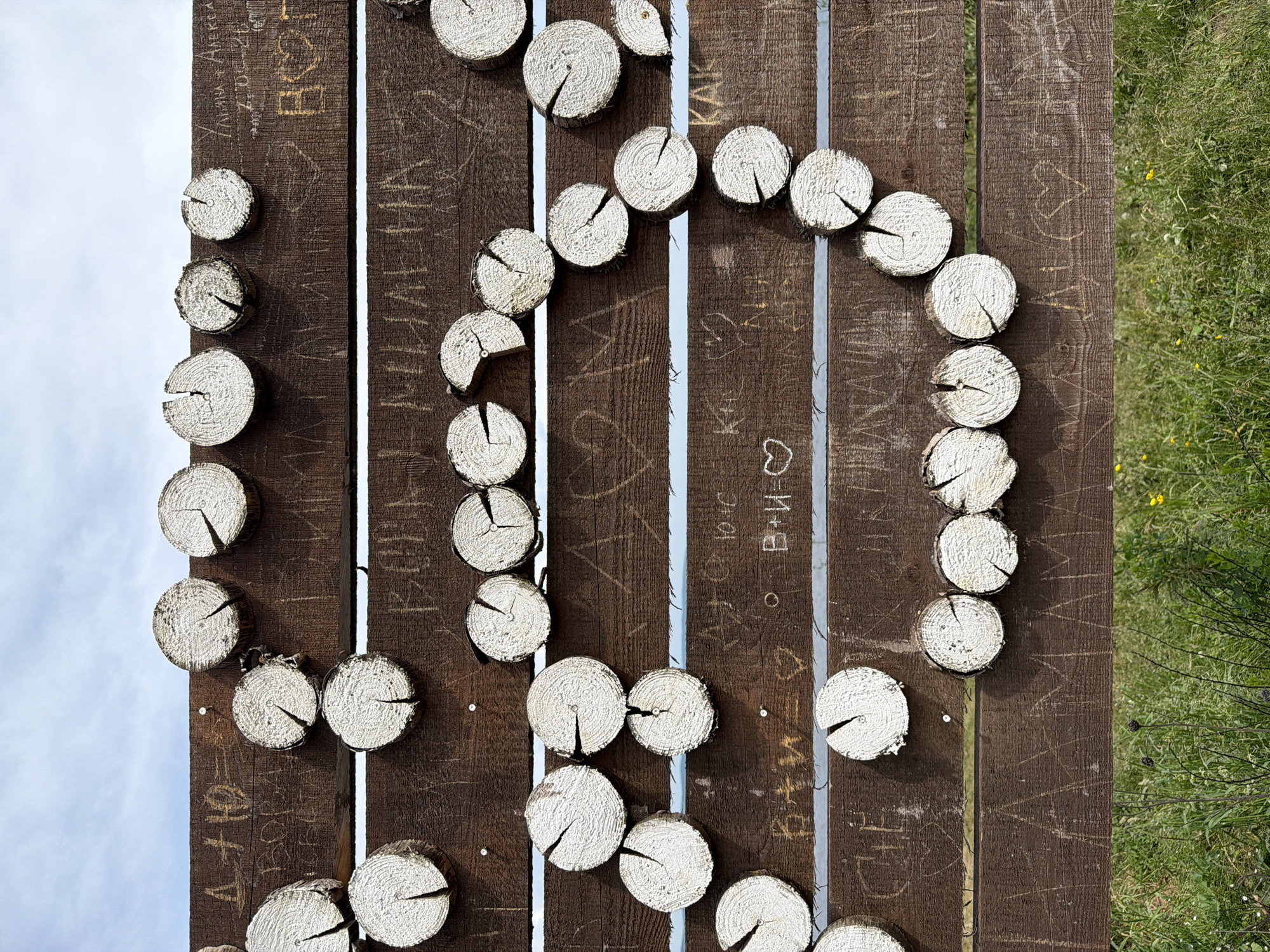 The image depicts a rustic wooden fence with several rows of horizontal planks. Affixed to the planks are numerous circular wooden log slices, some of which are painted white on their visible faces, showcasing the wood grain and cracks. The fence is adorned with various inscriptions and carvings, including initials and symbols like hearts, suggesting it is a place where people leave messages or declarations. In the upper portion of the image, a cloudy sky is visible, while the lower portion shows a grassy area with some green vegetation and a few small yellow flowers, indicating an outdoor rural setting. The location is identified as Костоваты, Russia. There are no people or animals visible in the image. The scene is static and appears to be a permanent structure.