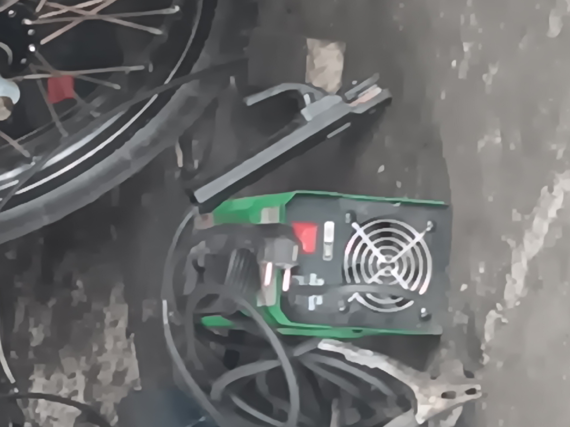 In Batursari, Indonesia, an image captures a scene with a green welding machine positioned on a concrete surface. The machine features a circular fan on its front panel and is connected to thick black cables that are coiled around it. To the right of the welding machine, a dark object with a metallic sheen and a gray stripe is visible, its purpose unclear from the image. In the background, the tire and spokes of a bicycle or motorcycle are prominent, with a hint of a red component on the wheel. The ground beneath the objects appears to be a rough, gray concrete or asphalt surface, with some scattered debris. The overall lighting suggests an outdoor setting, possibly in a workshop or yard.