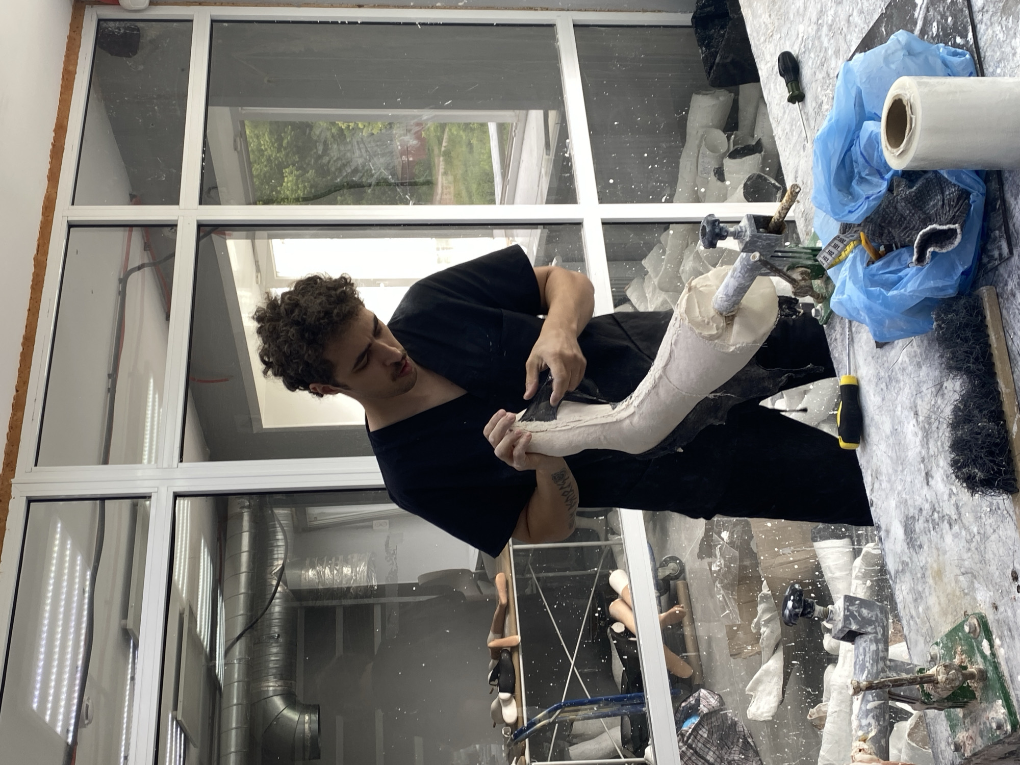 A young man in a dark blue scrub top and black pants is working on a prosthetic limb in what appears to be a workshop. He is holding the prosthetic with both hands and appears to be applying material to it with his right hand. The prosthetic is a pale color and has a textured surface. The man has curly brown hair and a light complexion. He is looking down at the prosthetic, focused on his work.
The workshop is filled with various materials and equipment related to prosthetics. In the background, through a large window, trees and greenery are visible, suggesting an outdoor environment. The room itself is well-lit, with fluorescent lights on the ceiling. There are shelves in the background with what look like unfinished prosthetic components. On the workbench in front of the man, there are various tools and supplies, including a blue plastic bag, a roll of white material, and what appears to be plaster or casting material covering the surface. The overall impression is one of a dedicated craftsman at work. There is no specific visual information to place this scene in Vladimir, Russia.