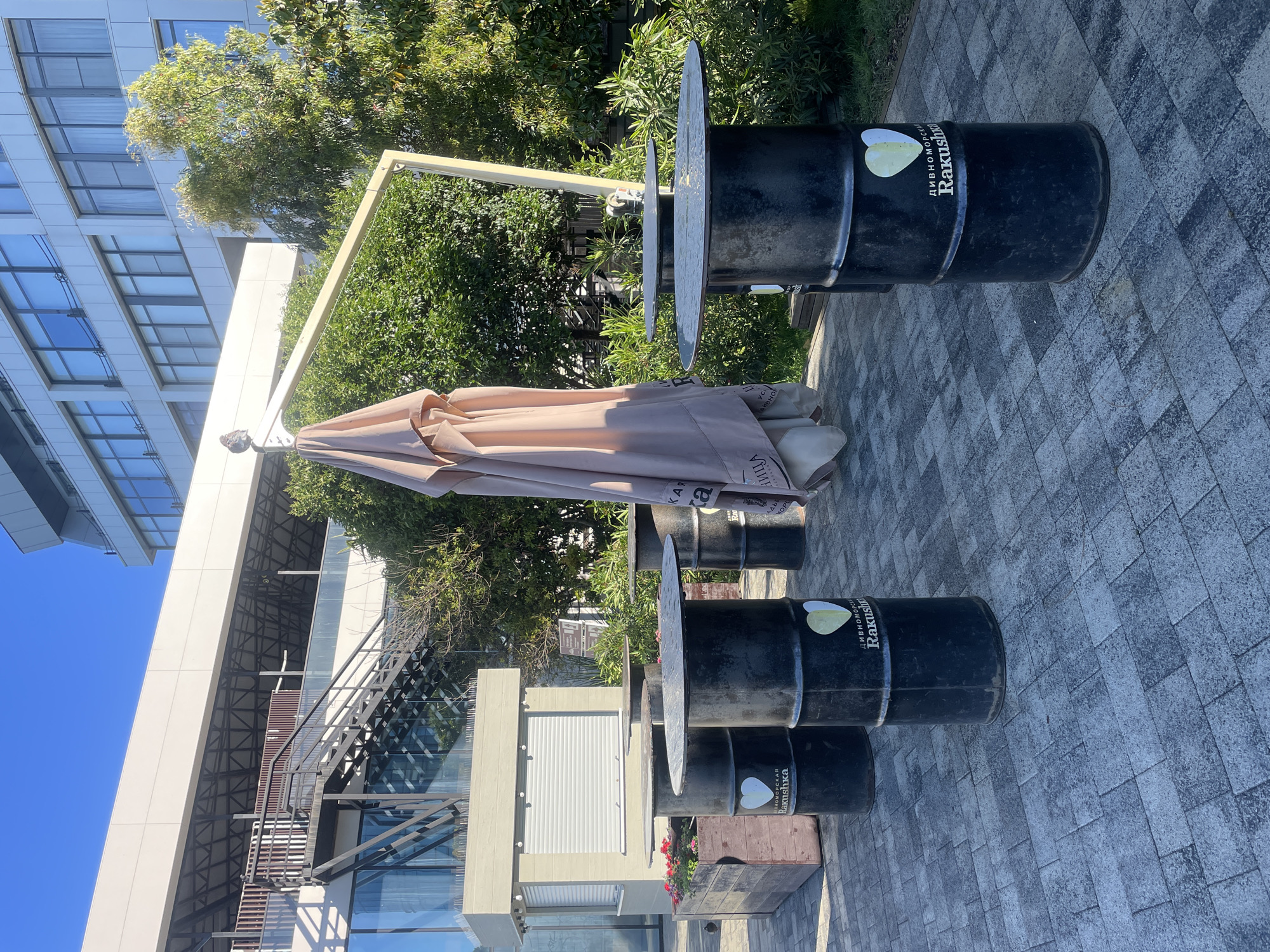 This image captures an outdoor patio area in Sochi, Russia, bathed in bright daylight under a clear blue sky. The scene features a series of black industrial-style barrels repurposed as tables, each topped with a round, flat metal surface. A large, folded light brown umbrella stands prominently in the center, suggesting a potential dining or relaxation spot.
The backdrop includes a modern multi-story building with a sleek, light-colored facade and numerous large glass windows, some reflecting the blue sky. An external metal staircase with dark railings is visible on the side of the building. Lush green trees and dense foliage are abundant, lining the perimeter of the paved patio area, which is covered in grey and black patterned tiles.
No people are present, indicating the area is currently unoccupied. The bright sunlight creates distinct shadows across the ground and objects. A wooden planter box filled with red flowers adds a touch of color near one of the barrel tables.
Visible text on the barrels includes "Rakushka" (Ракушка) in white lettering, accompanied by "дивноморское" (Divnomorskoe) and "пивного" (Pivnogo), suggesting a brand or establishment name, possibly related to a brewery or restaurant ("Divnomorskoe Beer Rakushka"). Each barrel also features a logo consisting of two overlapping, light-colored heart or pear shapes. The setting suggests an urban or commercial outdoor space designed for leisure, currently calm and unoccupied.