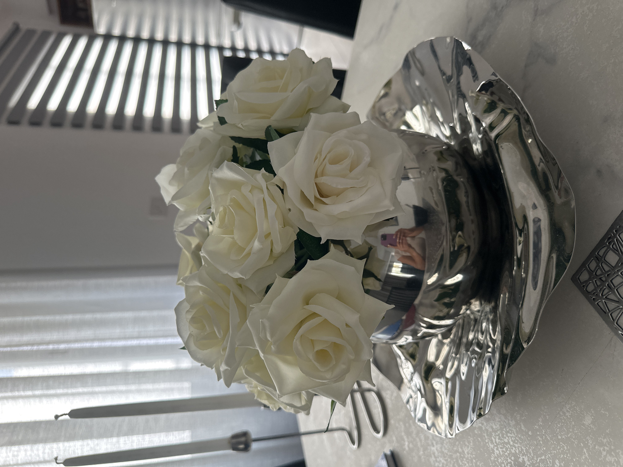 A close-up, eye-level shot shows a silver bowl filled with white roses on a silver decorative tray. The tray has a rippled, wavy edge. The arrangement sits on a light-colored, patterned surface, likely a dining table. In the reflection of the silver bowl, a person holding a pink phone is visible. In the background, sheer white curtains hang over a window, with two tall white candles in silver holders to the left. To the right, a window with horizontal blinds casts striped shadows across the room. The overall scene appears to be an indoor setting, likely a home.