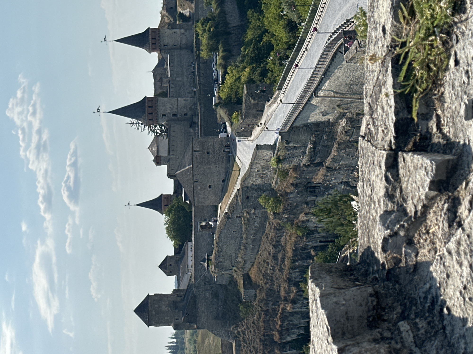 The image shows a view of the Kamianets-Podilskyi Castle in Ukraine. The castle is a large stone fortress with multiple towers, some of which have conical roofs with flags. A bridge with lampposts spans across a gap in front of the castle, leading to a road that curves up towards the fortress entrance. The castle is situated on a rocky, elevated terrain covered with dry vegetation and some green trees. The sky above is light blue with scattered white clouds. In the foreground, large rough-hewn stones are visible, suggesting the perspective is from another part of the fortification. There are a few people visible on the bridge and near the castle entrance. Cars are parked on a road near the castle walls. The overall scene depicts an ancient architectural landmark under a clear sky.