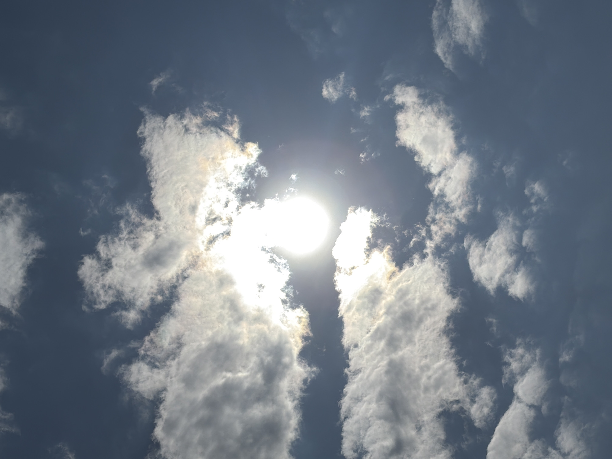 The image displays a bright, daytime sky dominated by the sun and clouds. The sun is a brilliant white orb, positioned centrally and slightly towards the top of the frame, casting a strong glare. It is partially obscured by various white and grey clouds that are arranged around it. Two prominent, elongated cloud formations stretch vertically on either side of the sun, creating a natural frame and allowing glimpses of the deeper blue-grey sky between them.
The clouds are varied in density and texture; some appear soft and wispy, while others are thicker and more opaque, with their edges brilliantly illuminated by the sun's light. Darker patches within the clouds suggest shadows and depth. The overall lighting indicates a clear, sunny day with significant cloud cover. No specific activity or event is taking place beyond the natural atmospheric conditions. There are no visible people, man-made objects, or discernible text within the image. The scene depicts a sky view, with no ground features or horizon visible. The location context provided is Прикамский, Russia.