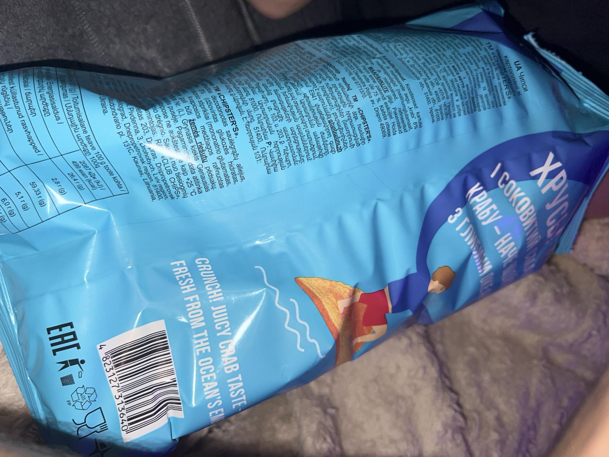 The image shows a close-up of a bright blue snack bag with prominent white text and graphics. The text is in several languages, including Ukrainian, English, and what appears to be Armenian. The English text reads "CRUNCH! JUICY CRAB TASTE FRESH FROM THE OCEAN'S EM". There is a graphic of a person on a surfboard and stylized waves. A barcode is visible on the lower right side of the bag, along with recycling symbols and the EAC mark.
The bag is lying on a textured, dark gray surface, likely fabric, which appears to be a blanket or similar material. The lighting suggests an indoor environment. There is no visible indication of the city of Zarichchia, Ukraine, in the image itself, nor any direct evidence of it being taken there. The information about Zarichchia is external to the visual content of the image.