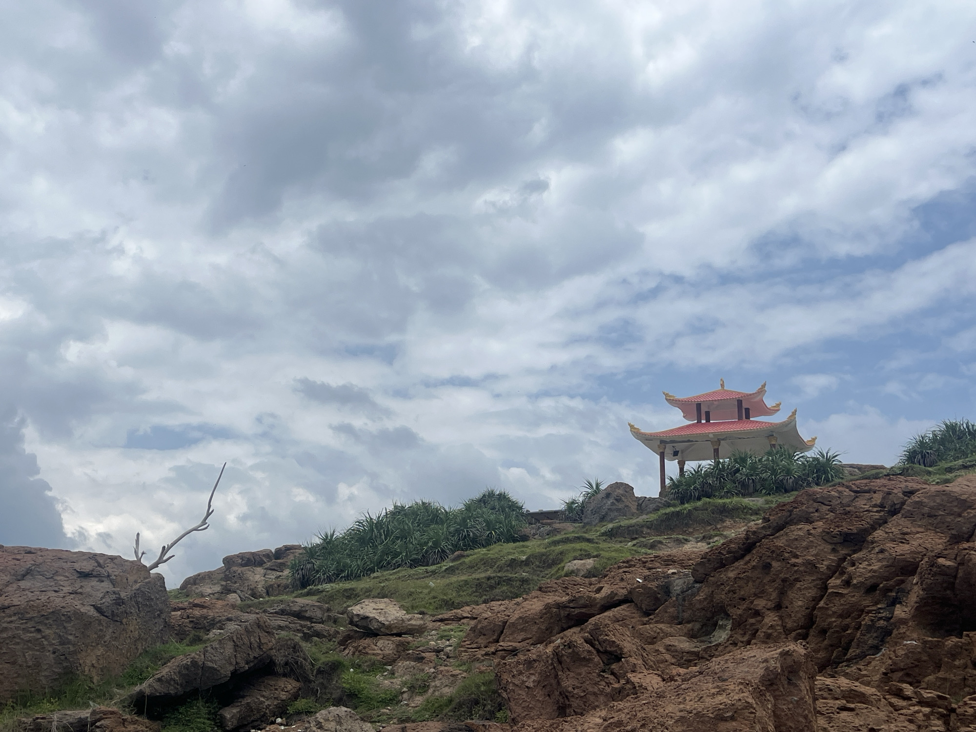 A pagoda-style building with a red roof and white walls sits on a grassy hill overlooking a rocky coastline under a cloudy sky. The building has multiple tiers with curved eaves, characteristic of East Asian architecture. Lush green vegetation, including spiky plants, grows on the slopes. Large, reddish-brown rocks are scattered across the foreground and midground, with a bare, twisted tree branch visible on the left. The sky is filled with dramatic, grey and white clouds, with patches of blue peeking through. The scene suggests a scenic overlook or temple in a coastal area, likely Mui Ne, Vietnam, known for its beaches and unique geological formations. There are no people or animals visible in the image.