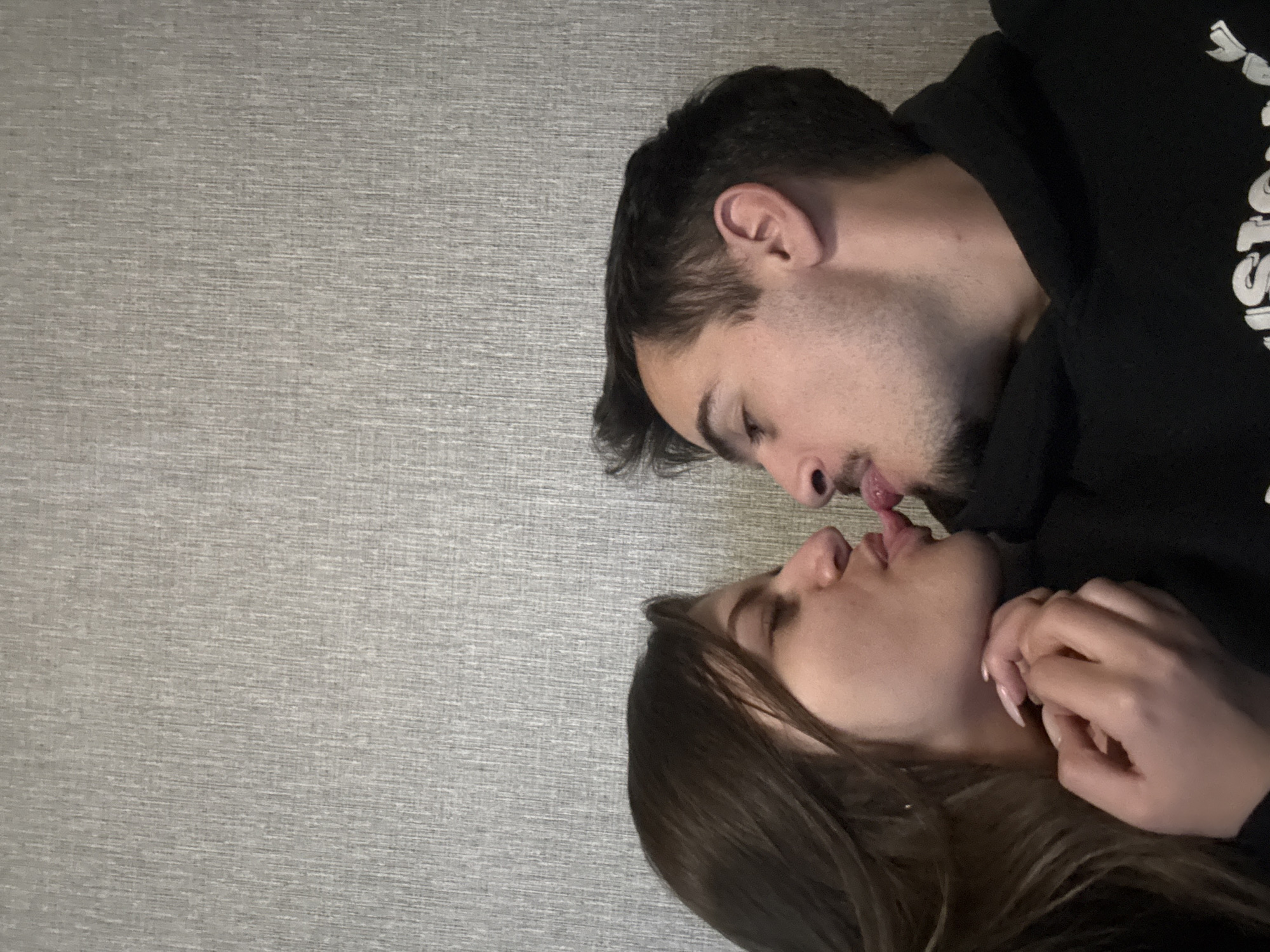 A man and a woman are shown from a high-angle perspective, their faces close together as if they are kissing. The man is wearing a black hoodie, while the woman has long, dark hair. They are lying on what appears to be a textured gray surface. The location is unknown but presumed to be in Sofia, Bulgaria.