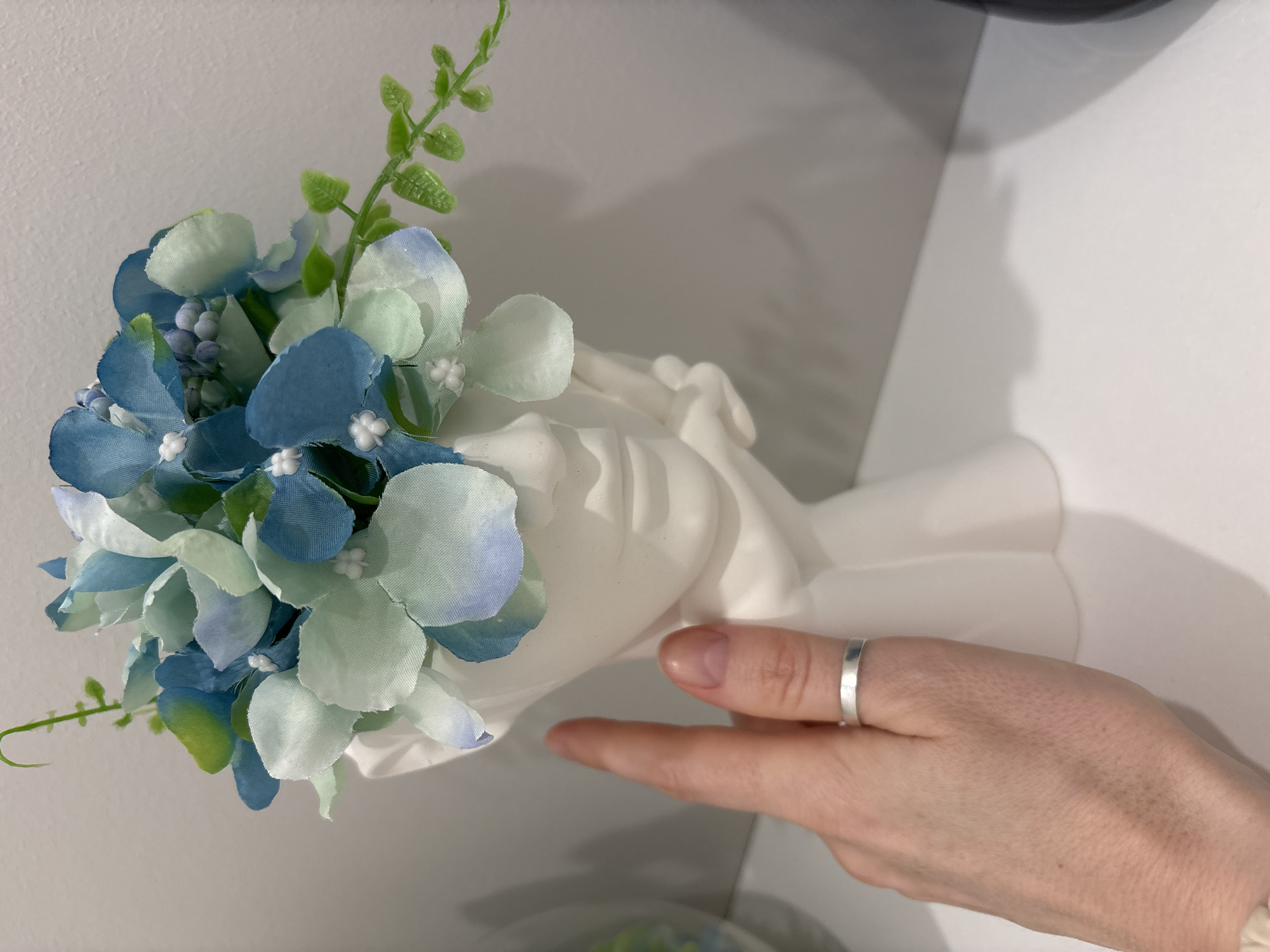 A close-up shot shows a white, hand-shaped vase filled with faux blue and green hydrangea flowers and small green foliage, held by a person's hand. The hand wearing a silver ring has the index finger pointing towards the vase. The background is a blurred white wall and countertop. There is no verifiable evidence to suggest that this image was taken in Saint Petersburg, Russia.