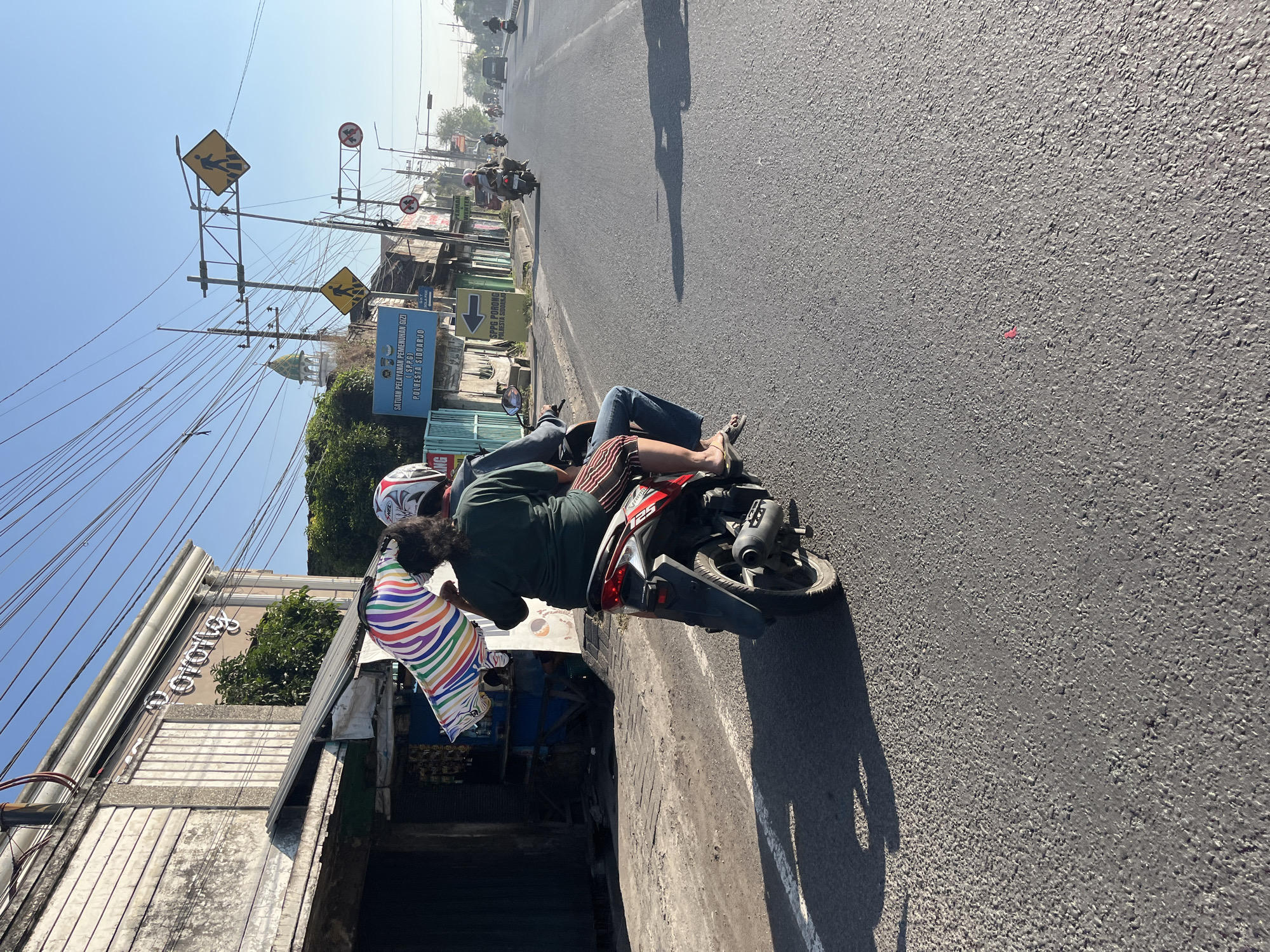A medium shot captures two individuals riding on a red and black scooter on a paved road in Siring, Indonesia. The rider in front, wearing a dark green shirt and striped pants, is seated behind the driver. The driver is wearing a blue jacket and jeans, with a white helmet. They are carrying a colorful, striped object, possibly an umbrella or banner, on their left. In the background, a white building with "Pusat Perbelanjaan" written on it is visible, along with a mosque minaret. The road is lined with shops and utility poles with many wires. Other vehicles, including motorcycles and cars, are present on the road, and shadows of the scooter and trees are cast on the pavement. The sky is clear and blue, suggesting it is daytime.