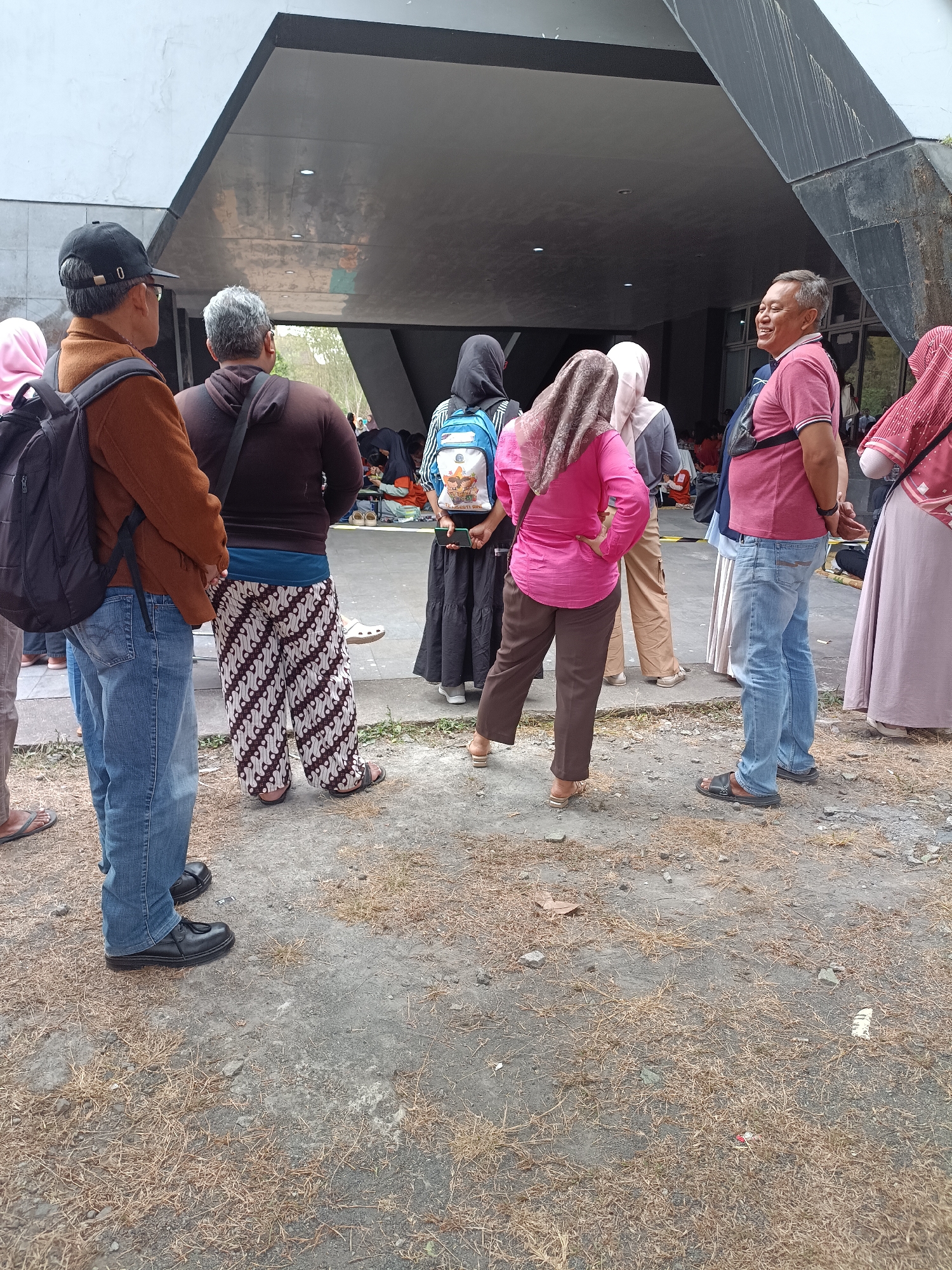 In Hargobinangun, Indonesia, a group of people are gathered under an overhang of a modern concrete structure. The scene appears to be outdoors, with dry grass and gravel covering the ground.
In the foreground, a man wearing a brown jacket, blue jeans, and black shoes stands facing away from the camera, a black backpack slung over his shoulder. To his right, another man in a brown hoodie and patterned pants also faces away. A woman in a bright pink shirt and brown pants is positioned with her back to the camera, her hands on her hips.
Further back, a man in a pink polo shirt and blue jeans is smiling and talking to someone off-camera. Several other individuals, mostly women wearing headscarves and long skirts or dresses, are visible in the mid-ground and background. Some are seated on the ground, while others are standing. One person is holding a smartphone. The overall atmosphere suggests a casual gathering or waiting period in a semi-public space.