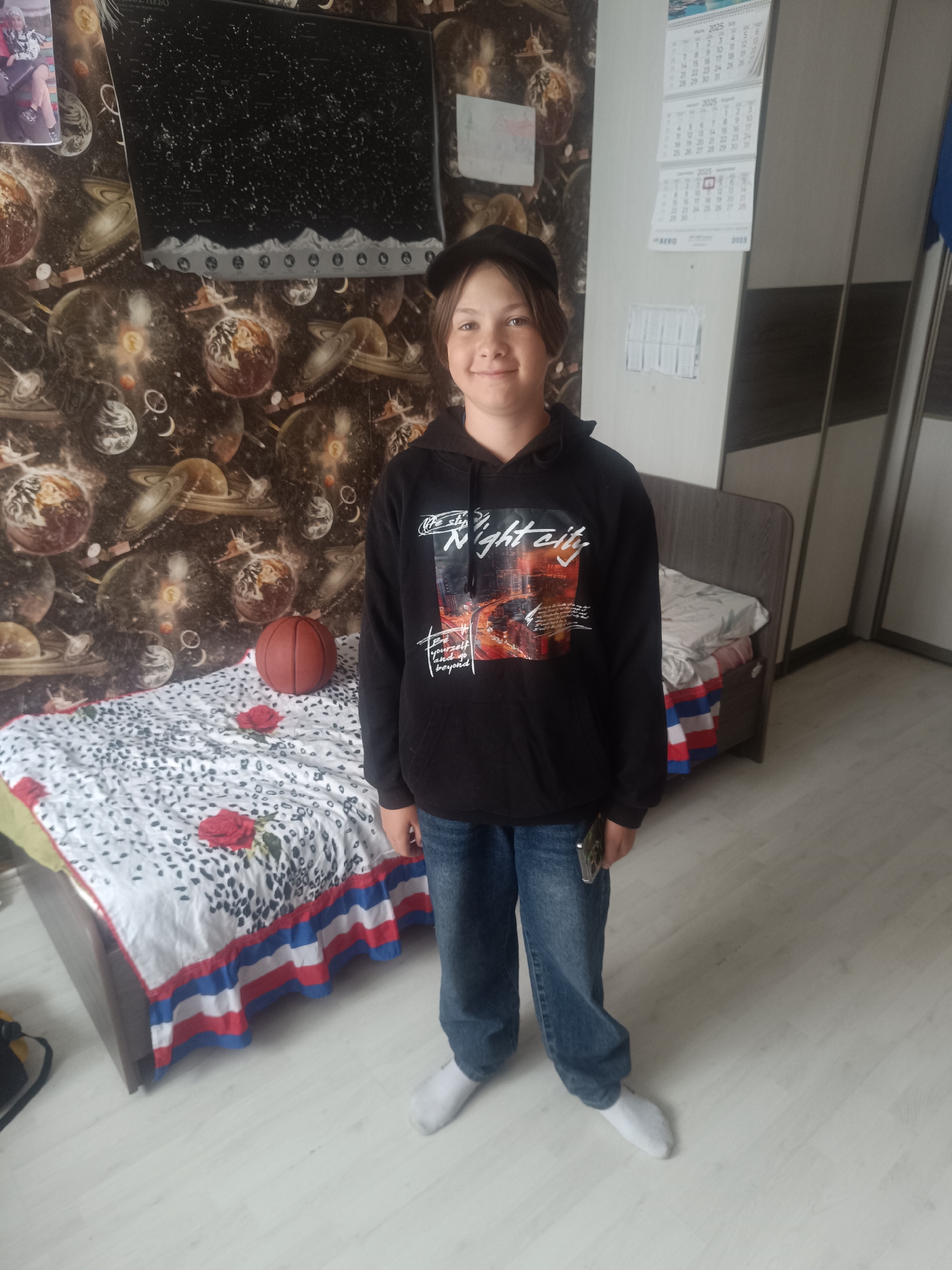 A young person stands in a room. The person wears a black hoodie with a graphic, blue jeans, and white socks, also wearing a dark colored cap. The room contains a bed with a white and black spotted comforter and red roses on top. A basketball is on the bed. The wallpaper is decorated with images of planets and space. A dark-colored TV or poster hangs on the wall, and a calendar is affixed to the wall near a closet. The floor is a light, neutral color. The location of the image is inferred to be within the city of Closed administrative–territorial formation "Mezhgorye", Russia, but nothing in the visual data suggests a location.