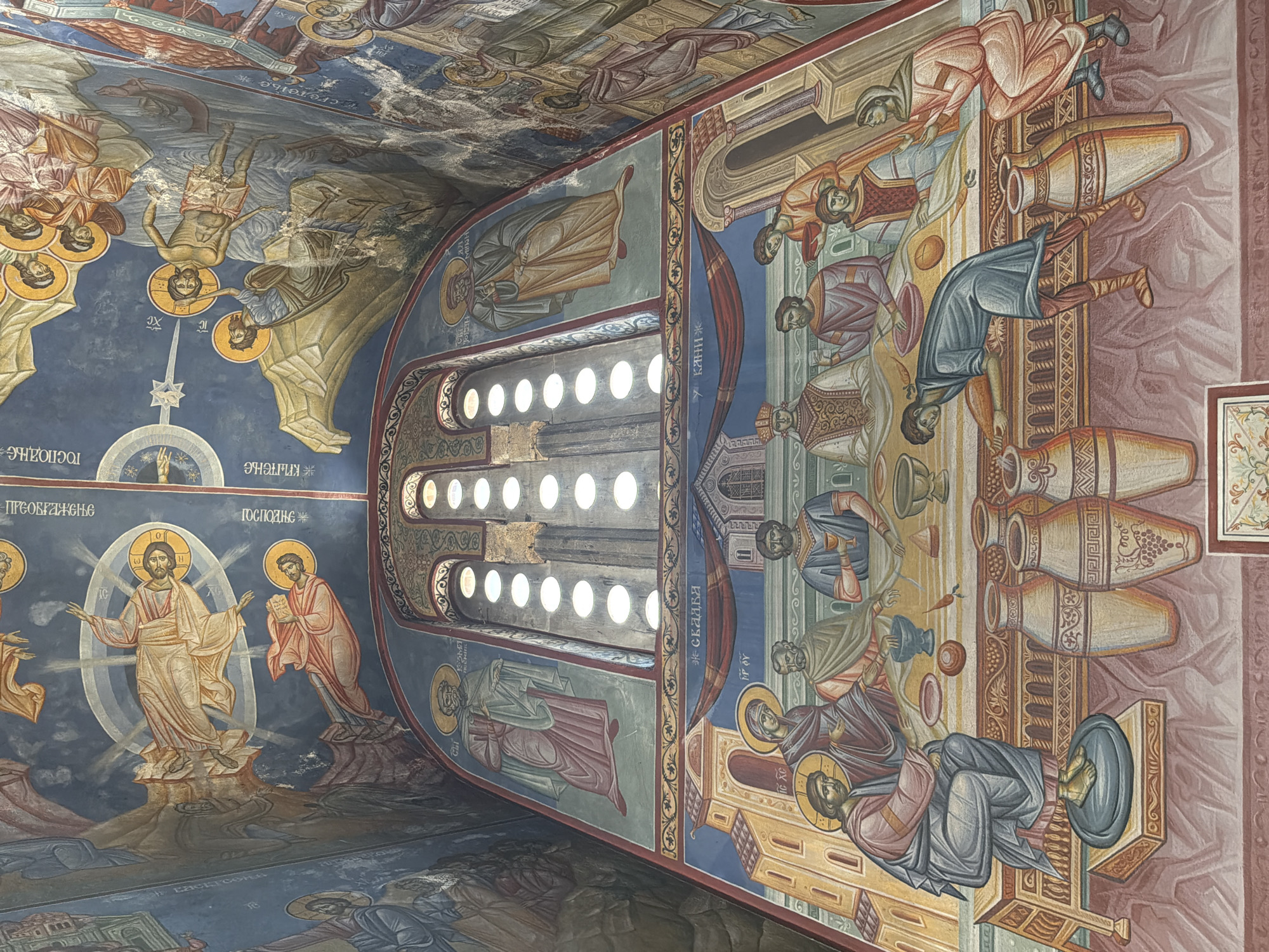 This image depicts a detailed fresco adorning the interior of the Church of the Holy Annunciation in Trebinje, Bosnia and Herzegovina. The fresco covers the ceiling and upper walls, depicting religious scenes in a traditional Byzantine style.
The central portion of the image focuses on a fresco above a large arched window. This fresco features figures in flowing robes against a deep blue background. In the upper section of the ceiling, Christ is depicted with a halo and surrounded by a circular mandorla. Below him, other figures with halos are visible, some in light-colored robes and others in red. The fresco continues with scenes of what appear to be biblical events, including figures being baptized or immersed in water, with rocky landscapes and starry backgrounds.
The lower section of the fresco showcases a narrative scene with multiple figures gathered around a table. A central figure, likely Christ, is seated on a raised platform, wearing a blue robe and with feet elevated. He appears to be interacting with individuals around the table, some of whom are seated and others standing. A prominent feature of this scene is a series of large ceramic vessels, resembling amphorae, arranged on the floor. Some of these vessels are intricately decorated with geometric patterns and grapevines. The figures are depicted with characteristic halo and stylized attire. The overall color palette is rich, with blues, reds, golds, and earthy tones. The environment is suggestive of an interior space within a church, with architectural elements and religious iconography.