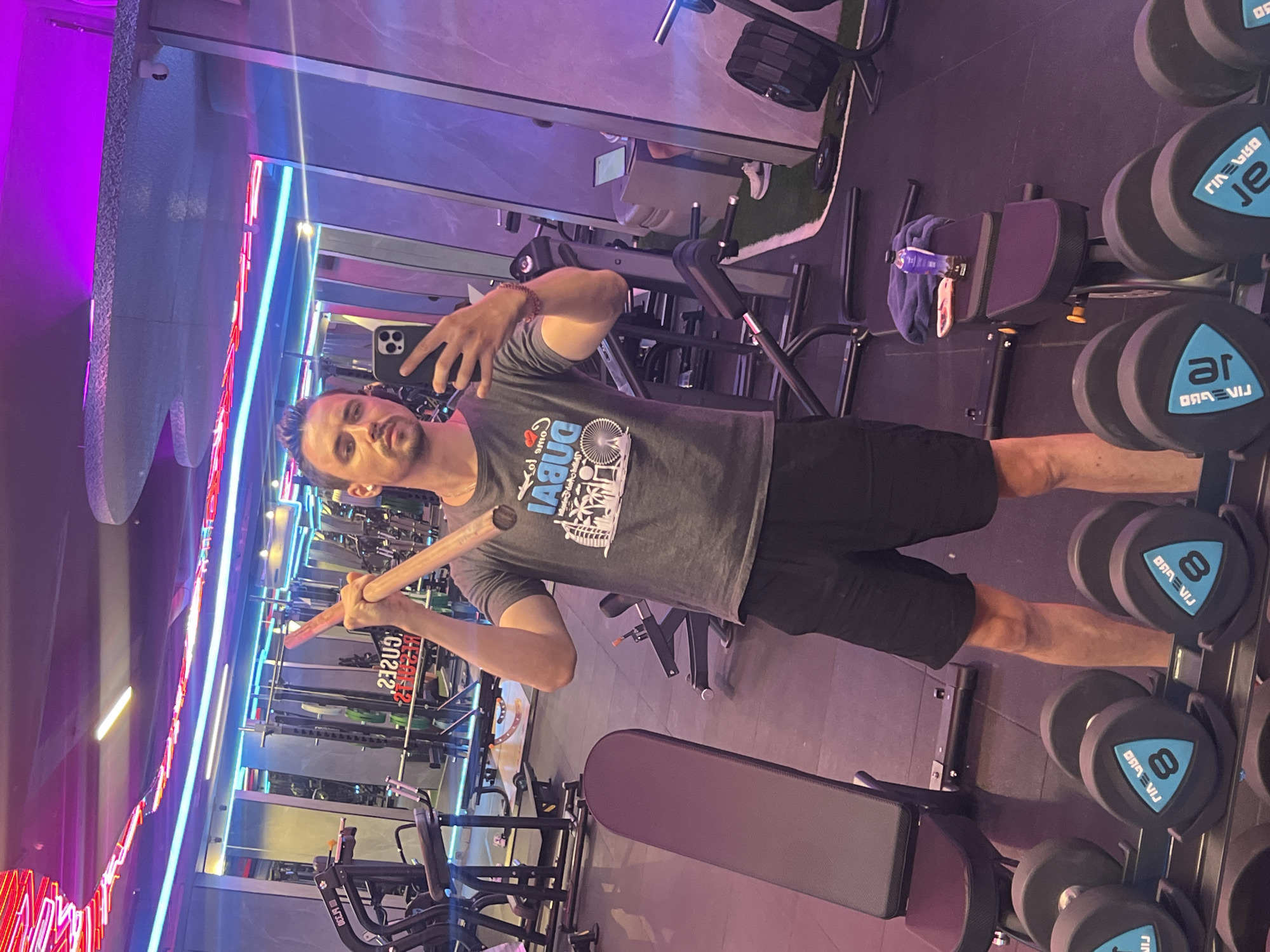 The image depicts a man taking a mirror selfie in a modern gym. He is wearing a grey t-shirt with a graphic print and black shorts, and is holding a wooden stick above his head. The gym has a sleek design with neon lighting in pink and blue hues. Various exercise equipment, including weight racks with dumbbells, benches, and a treadmill, are visible in the background. A water bottle and a towel are placed on a bench. The flooring appears to be a combination of dark rubber and artificial grass. The man's reflection is prominent in the mirror, capturing his face and upper body. The scene is well-lit, highlighting the gym's contemporary aesthetic. The image does not provide any specific information about its location in North Kuta, Indonesia.