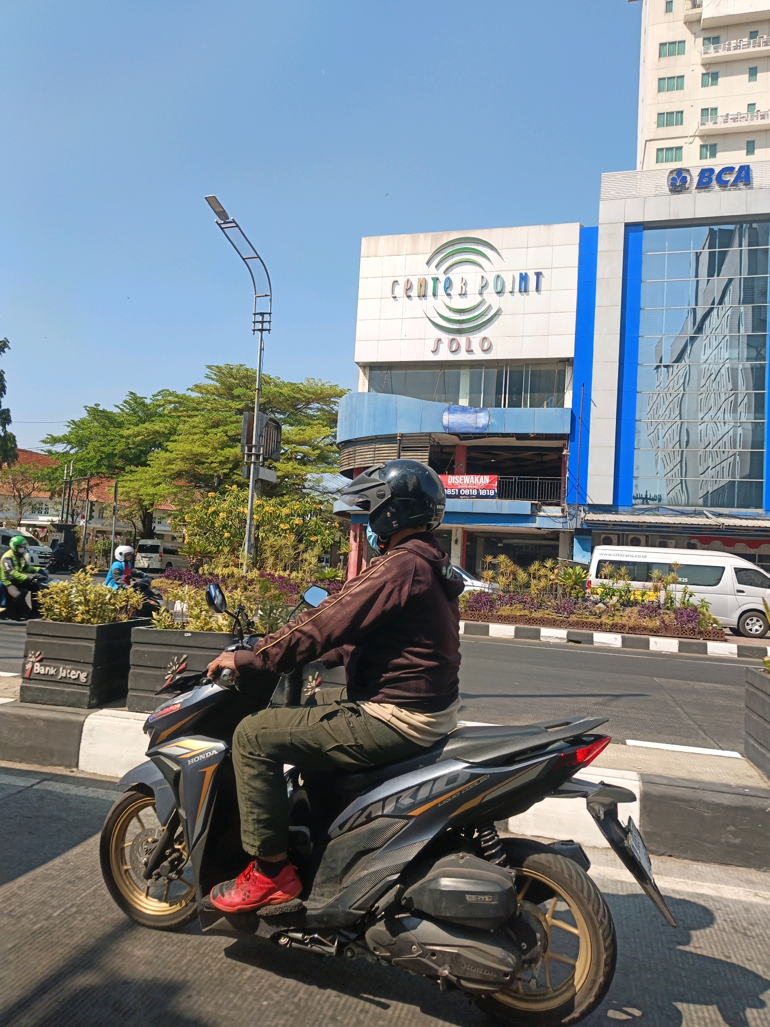 The image captures a street scene in Surakarta, Indonesia, on a bright, sunny day. In the foreground, a person wearing a brown long-sleeved shirt, green pants, a black helmet, and red shoes rides a black Honda motorcycle. The motorcycle has gold rims and displays the "Honda" logo. The rider is seen from the side, with their right hand on the handlebar.
Behind the motorcycle, a street with a clear lane is visible, with a gray and white striped median. Planters with green shrubs and purple flowers line the median. The planters have black signage that reads "Bank Jateng".
In the background, several buildings are present. The most prominent are "Center Point Solo," a white building with colorful signage, and a taller, modern white building with numerous windows, identified as "BCA" (Bank Central Asia) by its blue logo. Reflections of the sky and surrounding buildings are visible on the glass facade of the BCA building. Further back, trees with lush green foliage are abundant, and other buildings with red roofs can be seen.
The sky is a clear, light blue, indicating fair weather conditions. The overall environment suggests a busy urban setting with commercial establishments and traffic.