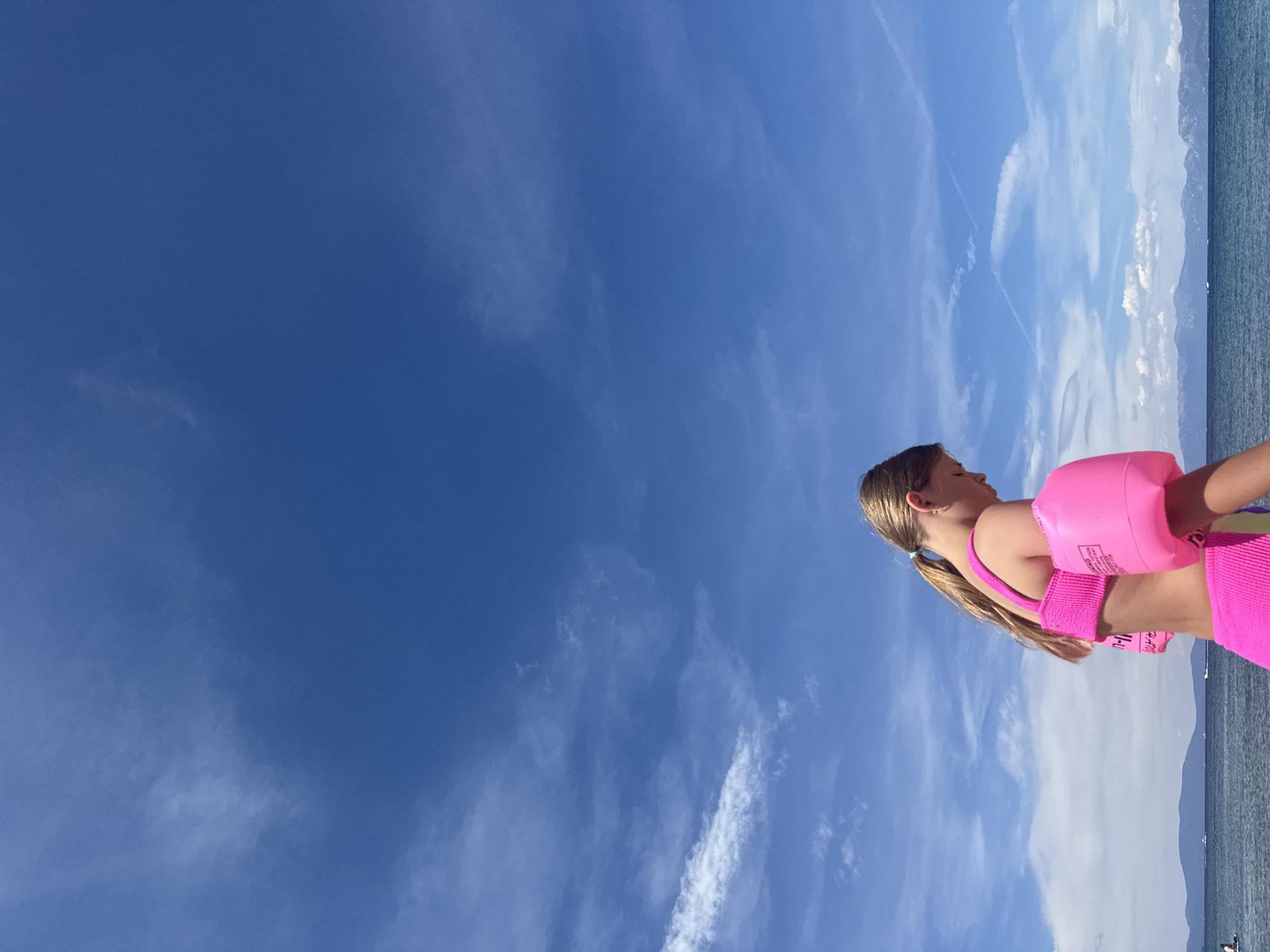A young girl, wearing a bright pink bikini and pink arm floaties, stands on a beach. Her hair is pulled back in a ponytail. The scene is set against a backdrop of a vast blue sky with scattered white clouds, and the calm, blue ocean stretches out to meet a distant, hazy shoreline with mountains. The sun appears to be bright, suggesting a clear day, possibly midday. The image is oriented vertically, with the girl positioned on the right side, looking out towards the ocean. There is some text visible on the arm floaties, but it is too small to be legible. The overall impression is one of a peaceful summer day at the beach, with the child likely preparing to swim or having just finished.