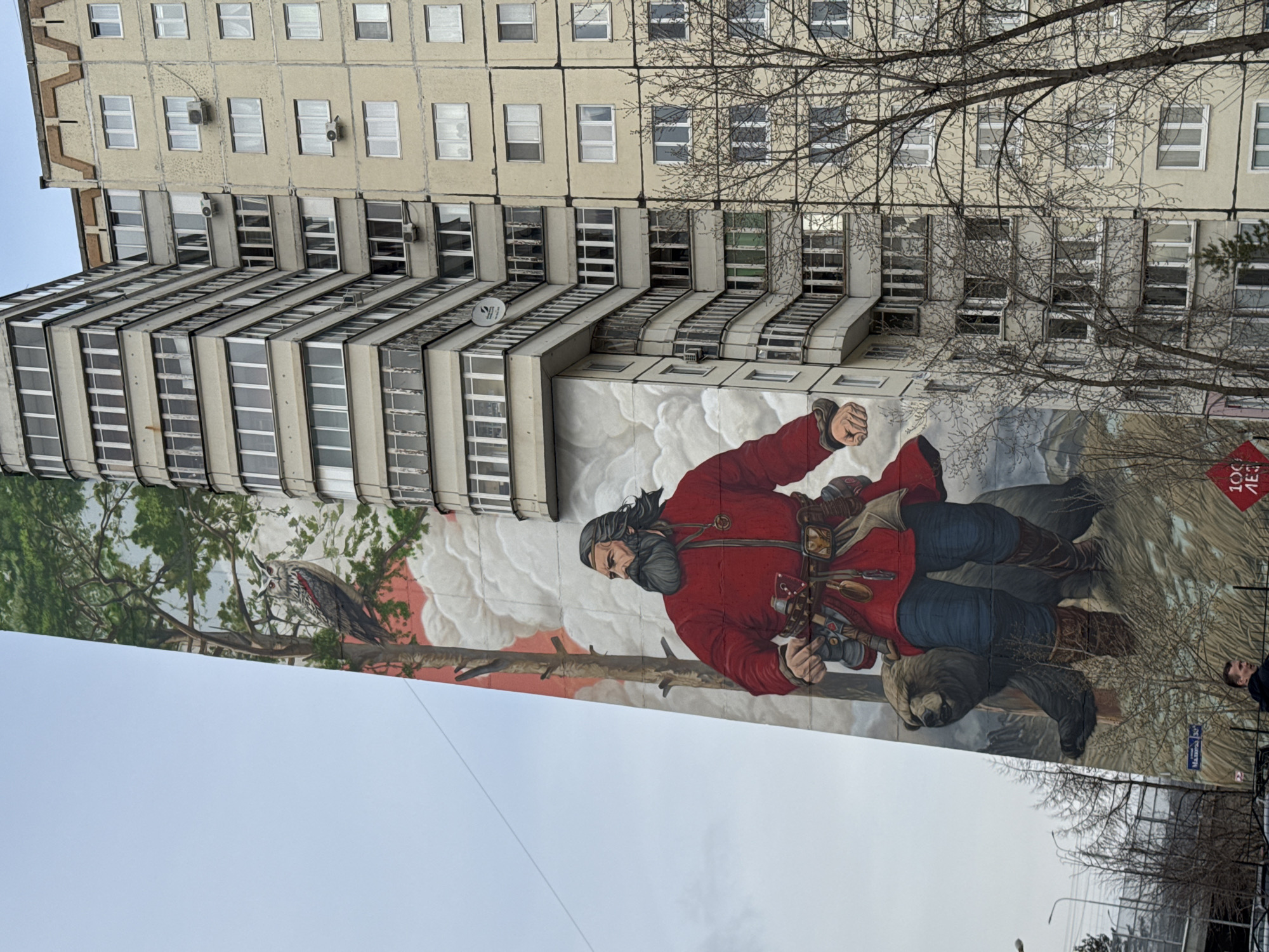 The image shows a mural painted on the side of a multi-story building in Perm, Russia. The mural depicts a man in a red coat, holding what appears to be a rifle or tool, with a bear and other natural elements depicted around him. The building is of Soviet-era construction, characterized by multiple rows of rectangular windows and balconies. Bare tree branches are visible in the foreground, partially obscuring the building. Street signs and a small sign that contains Russian writing are present at the base of the building. The sky is overcast.