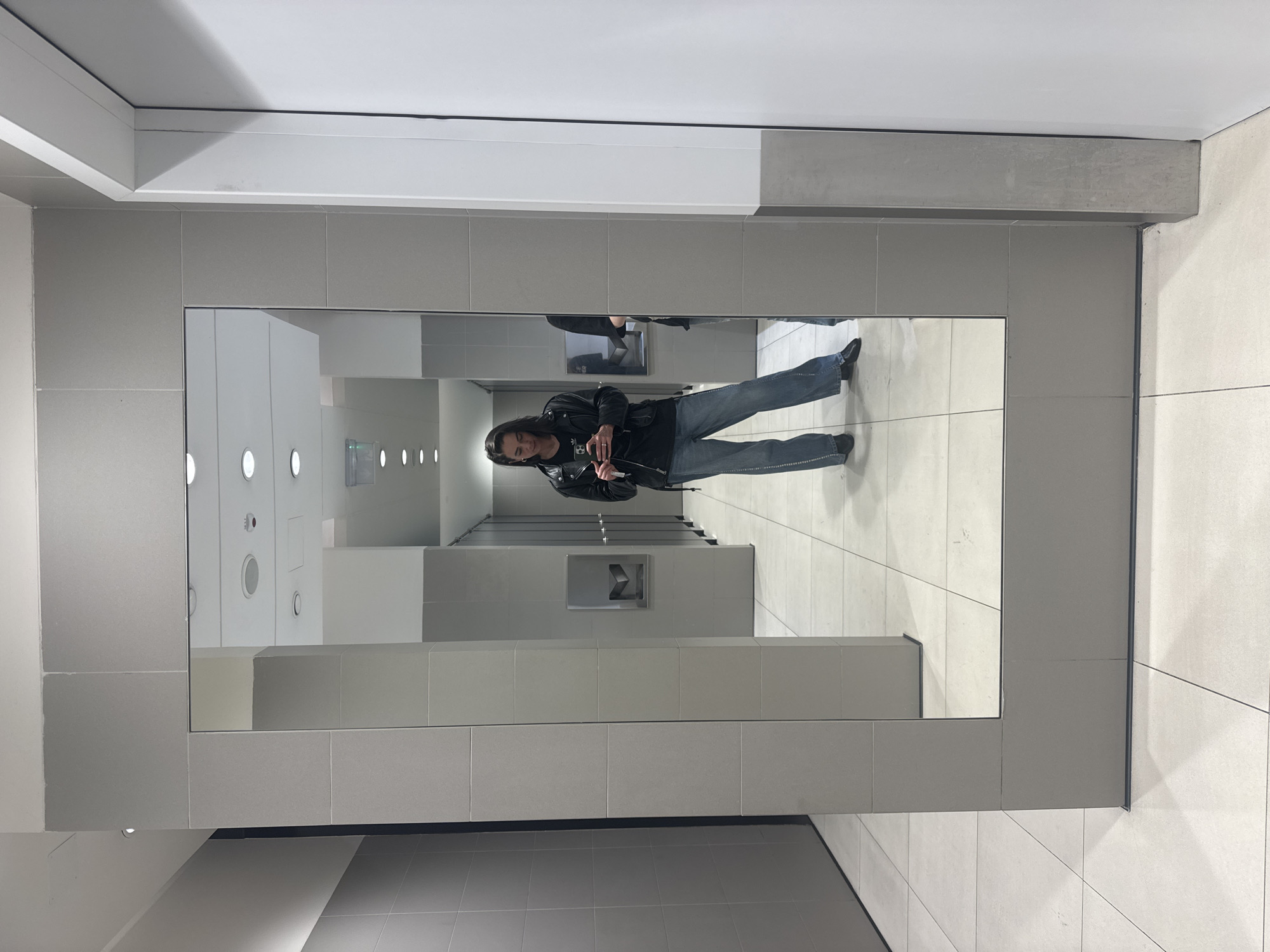A person in a black jacket and blue jeans is taking a selfie in a restroom mirror. The restroom has white and gray tiled walls and floors. The reflection in the mirror shows the interior of the restroom with multiple sinks and lighting fixtures. The location could potentially be in Belgrade, Serbia, but there is no specific information to confirm this.