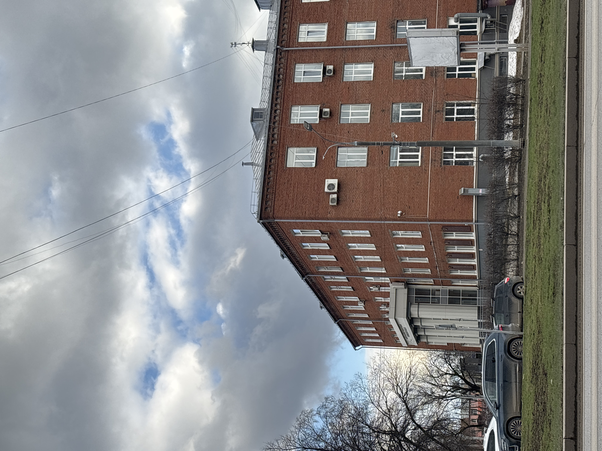 The image depicts a red brick building with multiple windows, some with air conditioning units, set against a sky filled with clouds. Overhead power lines stretch across the frame. A strip of green vegetation and a paved road run alongside the building, with several parked vehicles visible. Based on visual features and the style of architecture, the image may potentially show a building in Perm, Russia.