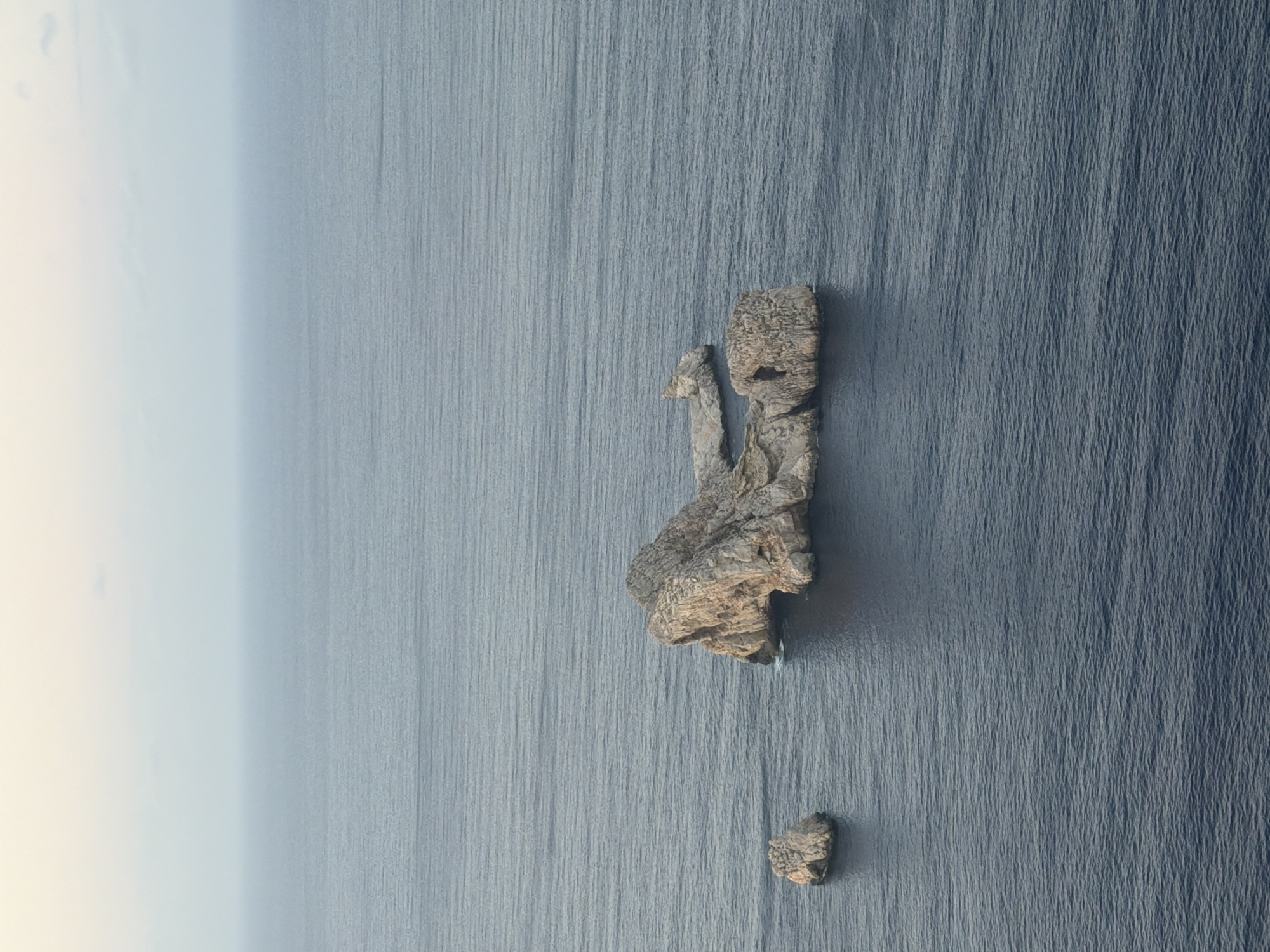The image is an aerial view of a rocky island formation in a dark blue sea. Two distinct rock formations are visible: a larger, more complex one with an archway, and a smaller, solitary rock to its left. The sea is calm with gentle ripples. The sky is a pale, hazy gradient of white and light blue, indicating either dawn or dusk. The scene is devoid of human presence or any structures. The geographical location is near Sant Antoni de Portmany, Spain, as suggested by the characteristic rock formations and seascape of the Balearic Islands.
