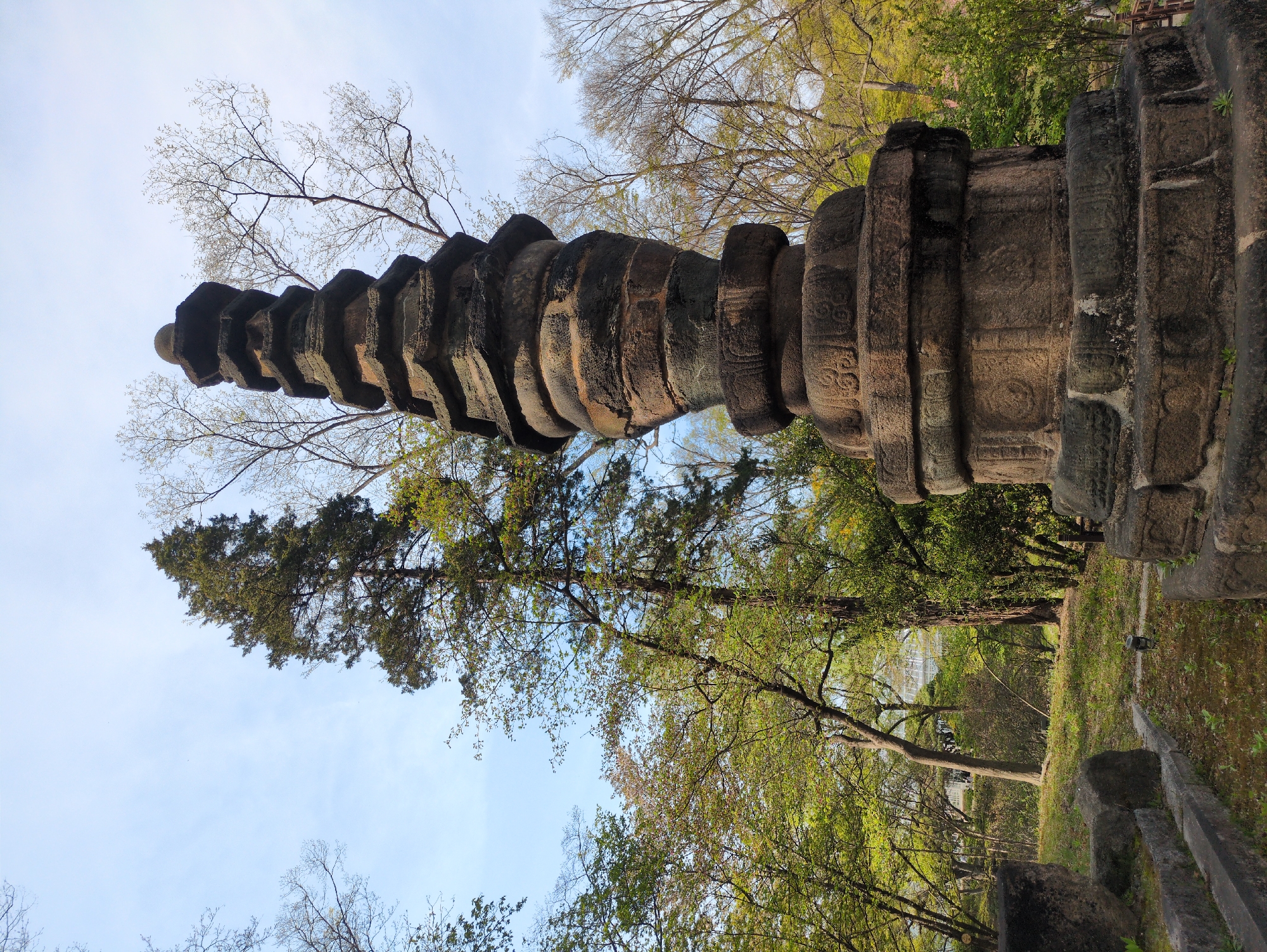 The image shows a stone pagoda in Seoul, South Korea. The pagoda is multi-tiered and made of dark stone. It is surrounded by trees with green foliage. The sky is light blue with some bare branches visible. The image appears to be taken from a low angle. The exact location in Seoul is not discernible from the image.