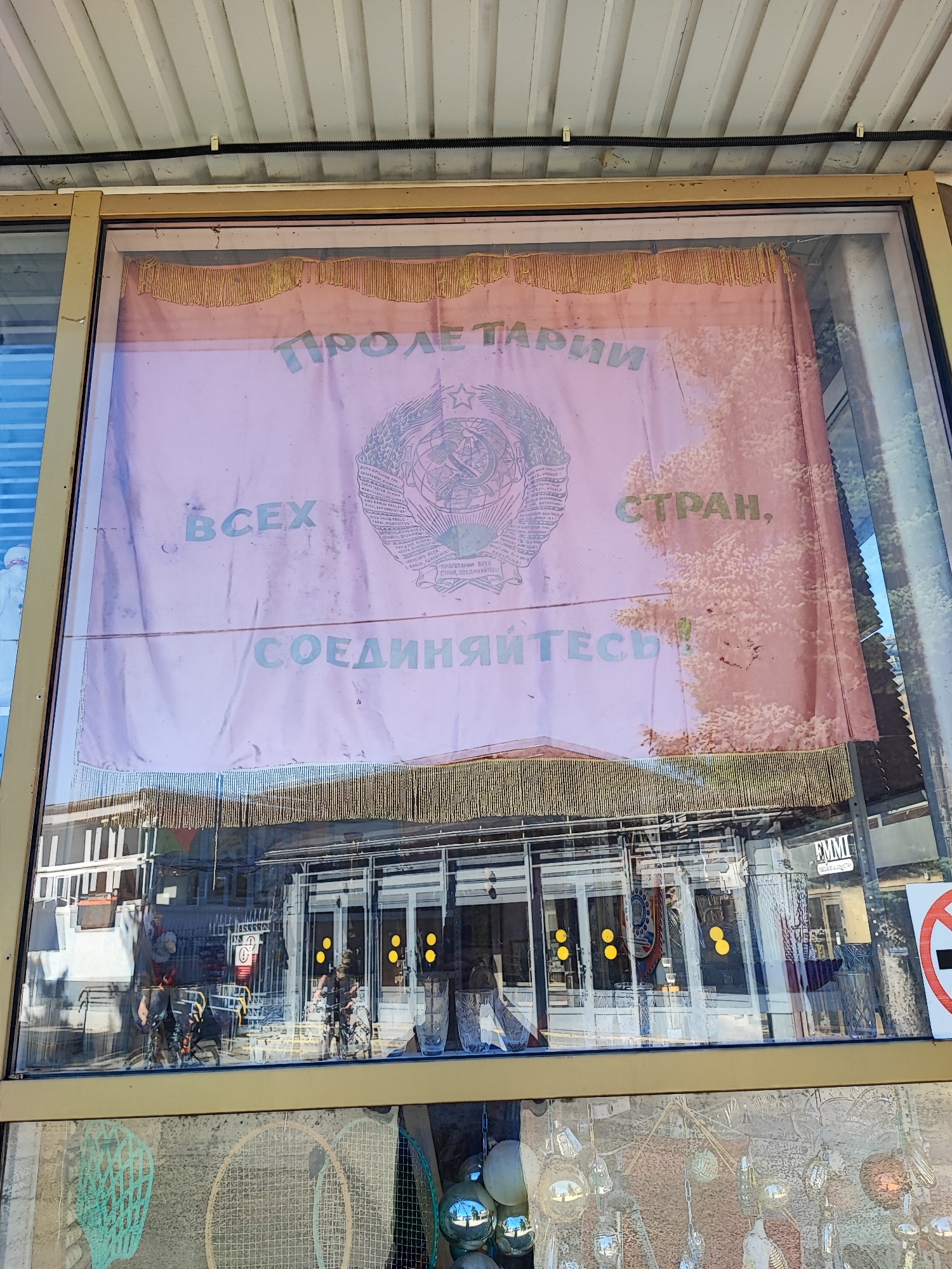 This image shows a window display in Pyatigorsk, Russia, featuring a red banner with Soviet-era iconography. The banner displays the phrase "Пролетарии всех стран, соединяйтесь!" which translates to "Workers of the world, unite!". This slogan is accompanied by the Soviet coat of arms, a hammer and sickle emblem surrounded by a wreath of wheat. The banner appears to be aged and slightly faded, with a fringe of gold tassels along the bottom.
Reflected in the window are buildings with glass doors and windows, suggesting a commercial area. Inside the establishment, various objects are visible, including glassware and what appear to be other display items. One reflection shows a sign with the acronym "EMMI" on it. Further below, another layer of the display is visible, showing what looks like decorative balls and items resembling tennis rackets, suggesting the establishment might be a shop selling various goods. The overall scene conveys a sense of historical display within a contemporary setting.