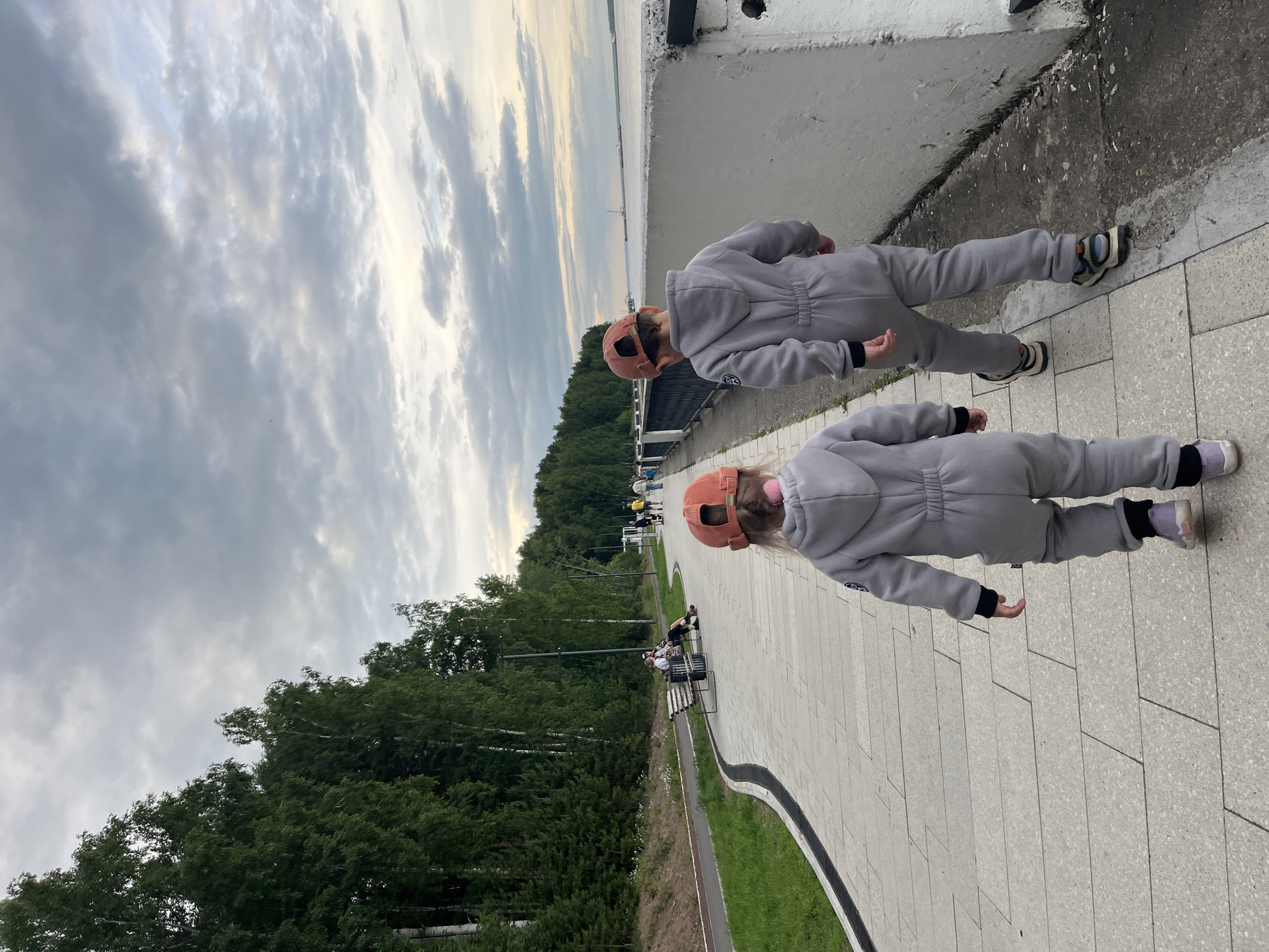 The image depicts two young children wearing grey hooded jumpsuits and baseball caps, walking along a paved walkway. The child in the foreground is wearing a peach-colored cap and the child behind is wearing a similar one. They are both facing away from the camera and walking towards the right side of the frame. Their shoes are visible, with the child in the front wearing purple and pink shoes and the child behind wearing white and blue sneakers.
The setting appears to be a park or waterfront area with a paved walkway lined with grass and trees. In the background, there is a body of water, possibly a river or lake, and a distant horizon with clouds. The sky is overcast with dramatic clouds, suggesting it might be late afternoon or evening, and possibly a bit windy. The overall mood seems casual and leisurely, with the children enjoying an outing.
There are other people visible in the background, some sitting on benches and others walking, indicating this is a public space. A railing or wall runs along the left side of the walkway. The path is made of light-colored paving stones, with a dark border along the edge. The trees are dense and green, creating a natural backdrop to the man-made walkway. No visible text is present in the image.