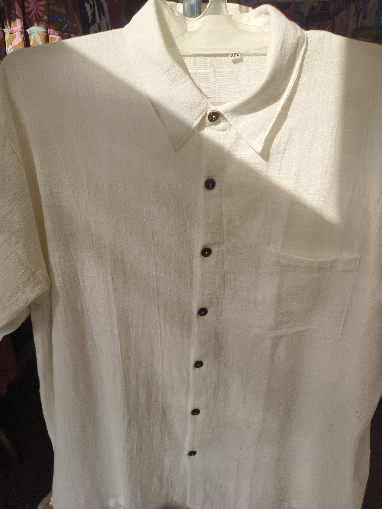 A white short-sleeved men's collared shirt is displayed on a hanger. The shirt is made of a textured linen-like fabric and features a button-down front with dark brown buttons. A single chest pocket is visible on the right side of the shirt. A size tag with "XXL" is attached to the inside of the collar. The shirt is hanging against a background of colorful fabric. A bright light casts a strong shadow across the upper portion of the shirt. The setting is a retail environment, likely a clothing store, in Kuta Selatan, Indonesia.