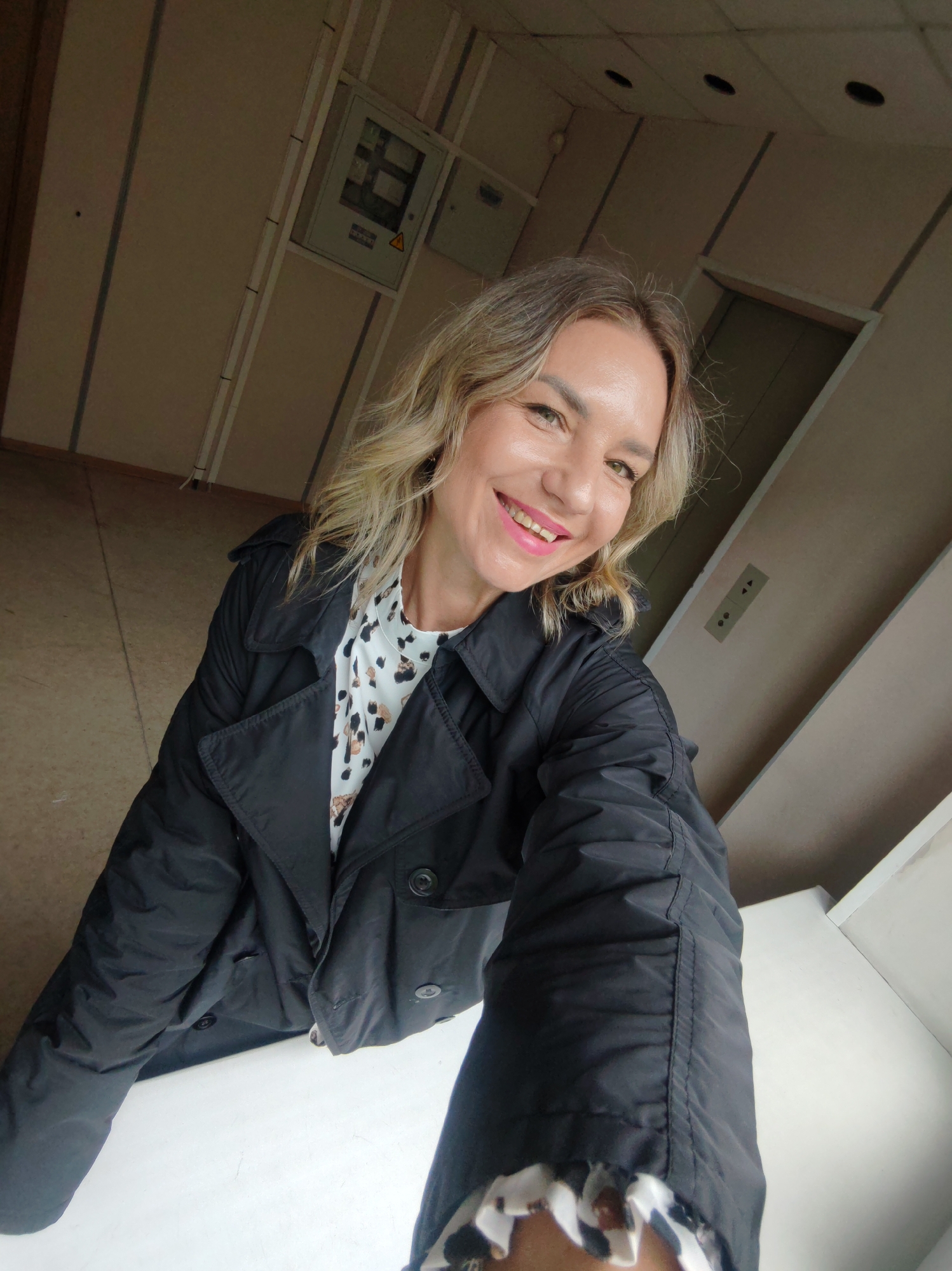 A woman is taking a selfie in what appears to be an office building, possibly in Petropavlovsk-Kamchatsky, Russia. She is wearing a black jacket over a printed top and has blonde hair. She is smiling broadly. In the background are walls painted in neutral tones, with an electrical panel and an elevator visible. The setting suggests an institutional or commercial space.