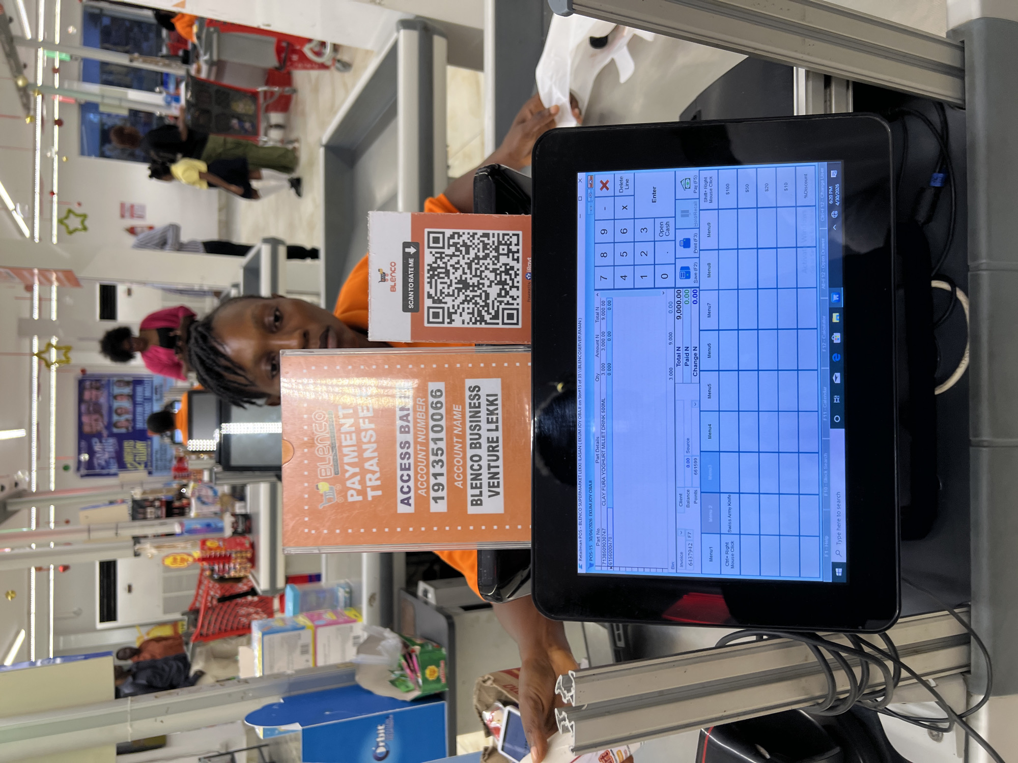 The image captures a cashier at a supermarket checkout counter in Igboefon, Nigeria, specifically in a Blenco Supermarket in Lekki. A person with dreadlocks, likely the cashier, looks towards the viewer while attending to the POS system. Several customers are visible in the background, some queuing and others moving within the store.
On the counter, two orange signs are prominent. One displays "BLEnco PAYMENT TRANSFER" with "ACCESS BANK" details, including Account Number "1913510066" and Account Name "BLENCO BUSINESS VENTURE LEKKI." The second sign features a QR code for "BLEnco SCAN TO RATE ME."
The POS terminal's touchscreen shows a transaction for "BLENCO SUPERMARKET LEKKI" dated "30/04/2026" at "6:20 PM." The screen details an item, "CLAY FURA YOGHURT MILLET DRINK 500ML," with a quantity of 3.000, totaling "N 9,000.00." The "Total N" is also "9,000.00," while "Paid N" and "Change N" are both "0.00," indicating an ongoing transaction. The interface includes a numeric keypad and options like "Open Cash," "Save (F2)," and "Pay (F5)."
The store interior is brightly lit, with supermarket shelves and products visible in the background. Festive star decorations hang from the ceiling. The scene depicts a routine retail transaction, highlighting available payment methods and customer feedback options in a modern supermarket setting.