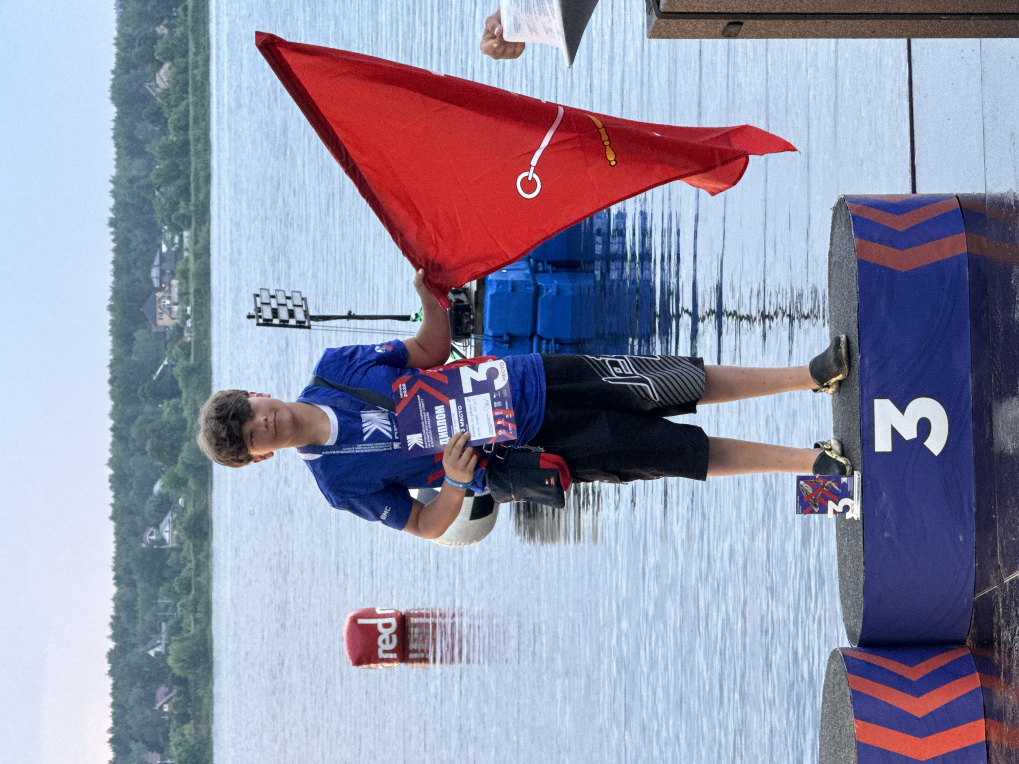A young male, wearing a blue t-shirt, black shorts, and dark shoes, stands on a podium. He is holding a red flag and a diploma. A soccer ball is tucked under his left arm. The podium has "3" prominently displayed. The background shows a body of water, trees, and buildings, suggesting an outdoor event. There is a red buoy with "red" written on it in the water to the left of the podium. In the distance, a white checkered structure with lights is visible. The sky is overcast. The diploma and bib indicate the event is a competition, and the number "3" suggests he placed third. The bib reads "ДИПЛОМ" (Diploma) and "3 МЕСТО" (3rd Place). The image appears to be taken at a waterfront location, potentially a competition held on a lake or river. While the image itself does not explicitly state the location is Perm, Russia, similar sporting events involving water and podiums are common in such areas.