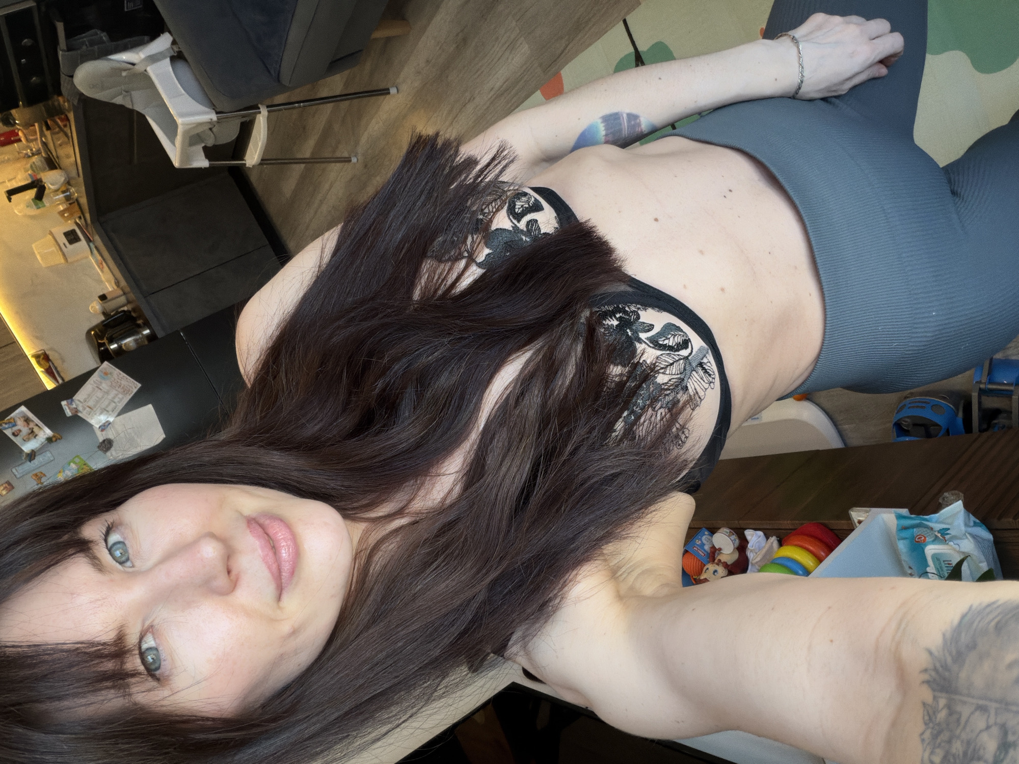 A woman lies on a floor in an indoor environment. She has long brown hair, fair skin, and blue eyes. She is wearing a black bra with lace detailing and gray leggings. Her right arm extends away from the camera, and she wears a bracelet on her wrist. A tattoo is visible on her upper right arm. The floor is made of wood-look vinyl plank. Furniture, including a high chair, a table, and a sofa, are visible in the background, along with children's toys and supplies. The scene is likely a domestic interior and there is no indication that it is in Perm, Russia.
