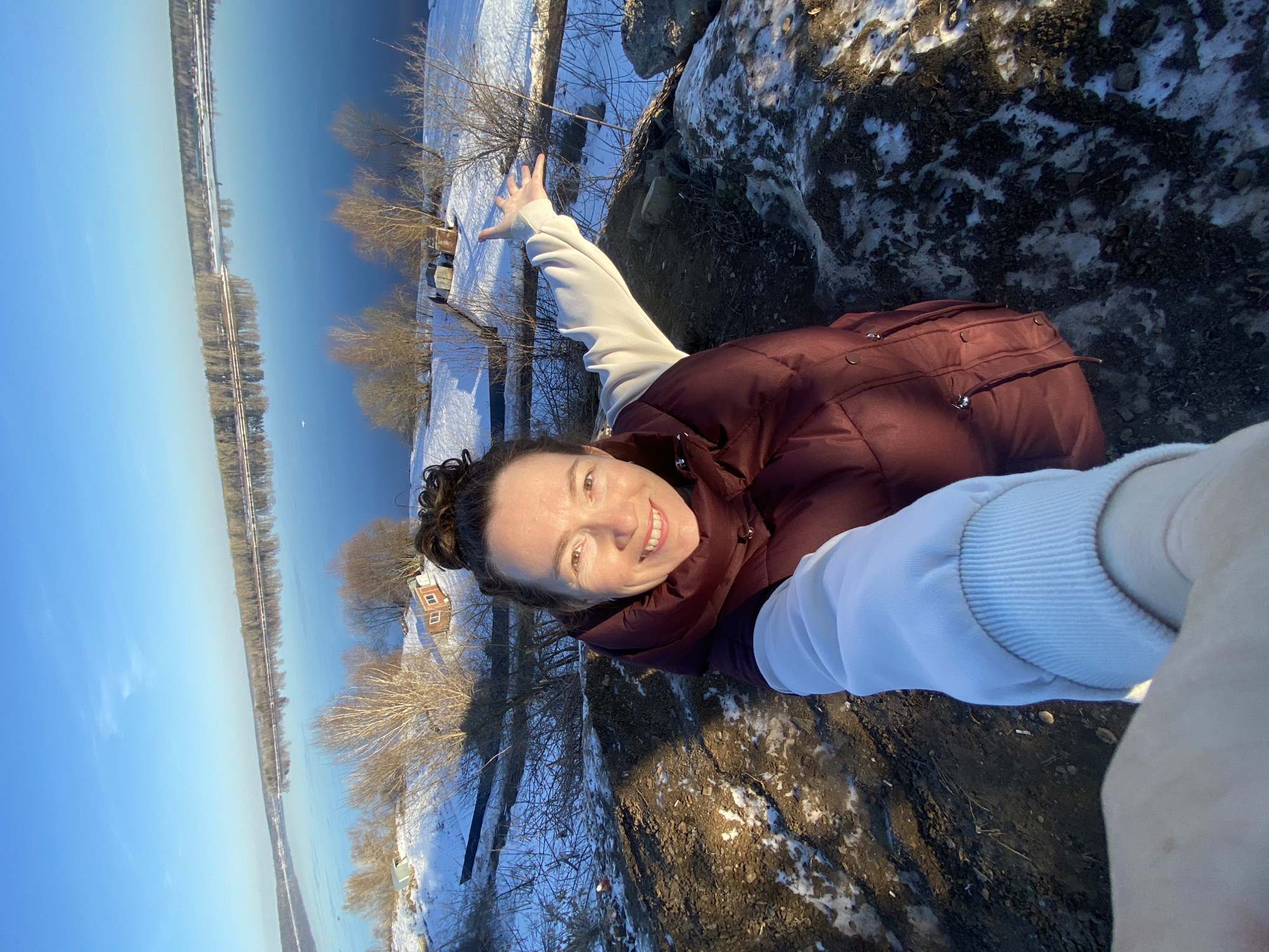 A medium shot captures a woman in Ольховка, Russia, as she stands on a snow-patched embankment near a river or large body of water. She wears a maroon vest over a light-colored sweatshirt and has her hair in a bun. She is smiling at the camera with her left hand extended towards the river. On the far side of the water are trees and buildings. The sky is blue. The embankment is composed of dirt and patches of snow.
